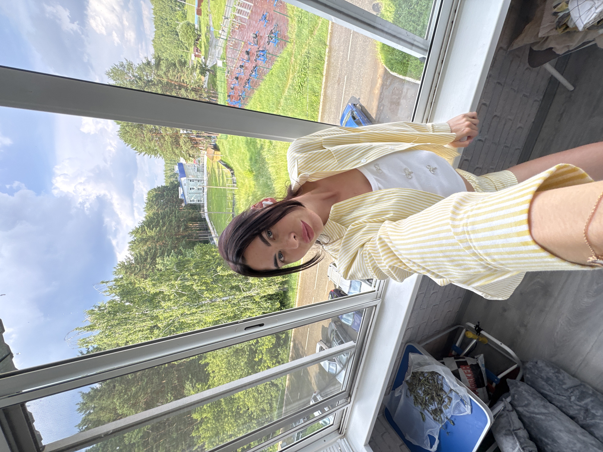 A woman with dark hair and light skin is taking a selfie from a sunroom with a view of a semi-urban, wooded area. She is wearing a yellow and white striped button-up shirt over a white tank top and matching shorts. The sunroom has large windows, offering a view of trees, a road with parked cars, a building, and an outdoor recreation area with exercise equipment. On a small table to the left, there is a plastic bag filled with dried plant material. The overall scene suggests a casual, possibly summer day in Chaikovsky, Russia.