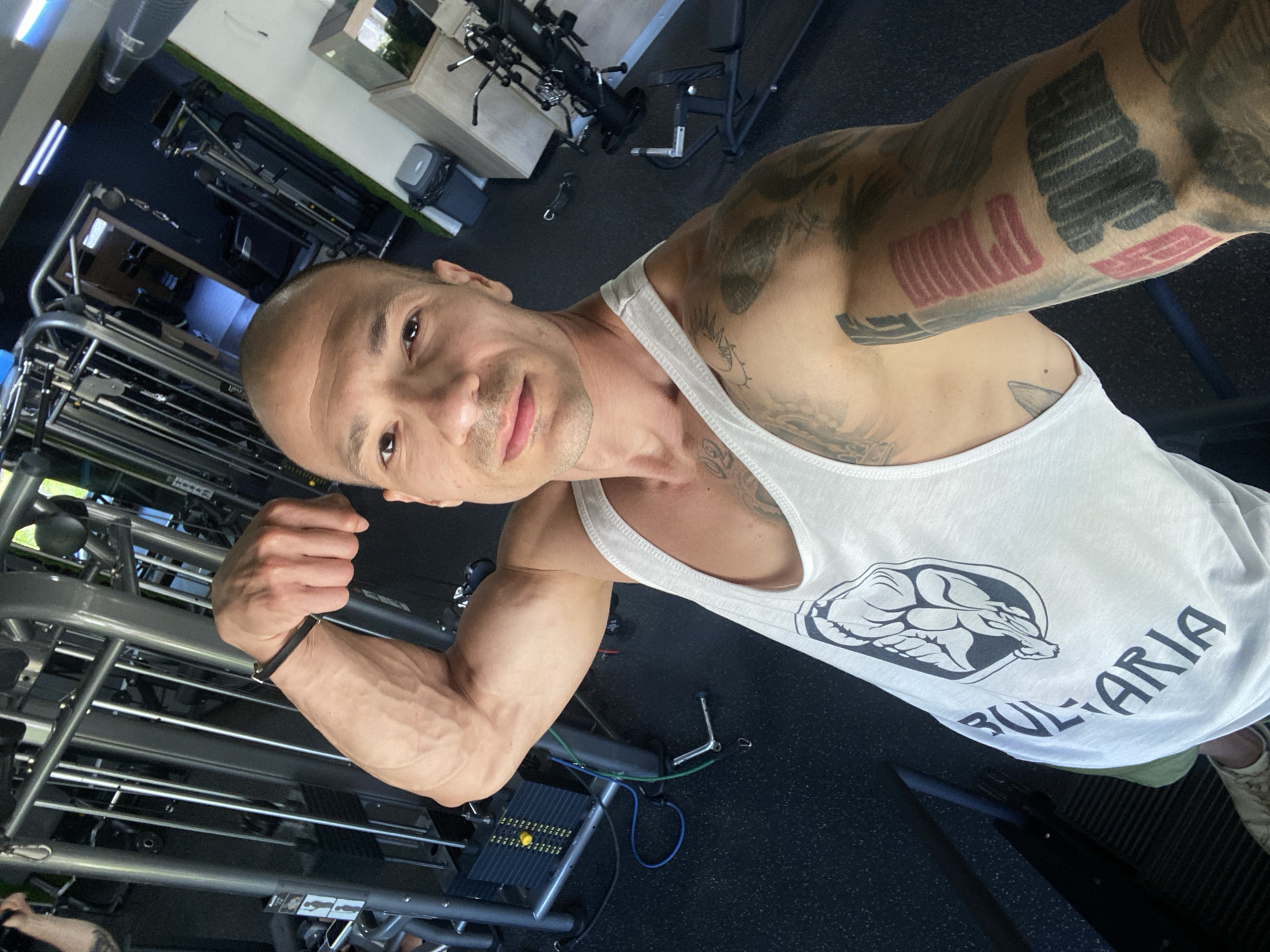 The image captures a man posing in a gym. He is wearing a white tank top with a graphic of a muscular figure and the word "BULGARIA" printed on it. He has tattoos on his arm. The gym appears to be equipped with various weight machines, and the flooring is dark rubber. The overall lighting suggests an indoor setting during the daytime. There is no specific indication of the city of Pleven, Bulgaria.