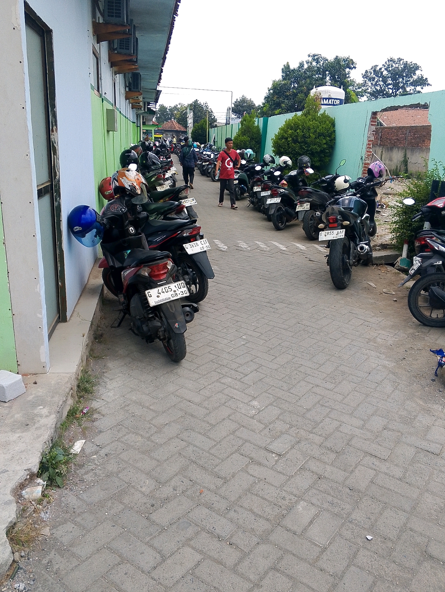 The image depicts an outdoor scene in Adiwerna, Indonesia, featuring a row of parked motorcycles lining a sidewalk adjacent to a building. The building has a light blue and green facade. Several motorcycles are parked in a line, some with helmets on them. A person in a red shirt is walking among the motorcycles. The ground is paved with gray interlocking bricks. In the background, there is a turquoise wall with a large white water tank on top, and trees. The overall atmosphere appears to be a parking area, possibly for a school or office, given the number of motorcycles.