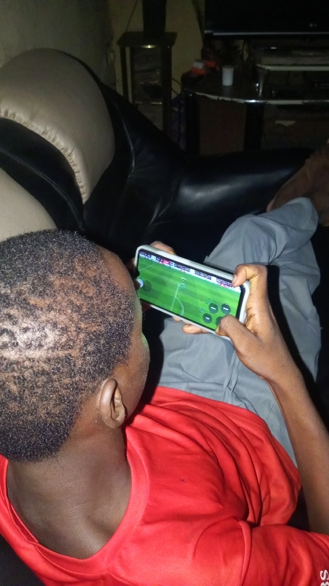 A person is lying on a black leather couch, wearing a red shirt and gray pants, holding a white smartphone in both hands and playing a soccer video game. The person has short, dark, curly hair. The environment includes a side table with a shelf underneath, and a television on a stand against a dark wall. The location is Unknown, Unknown.