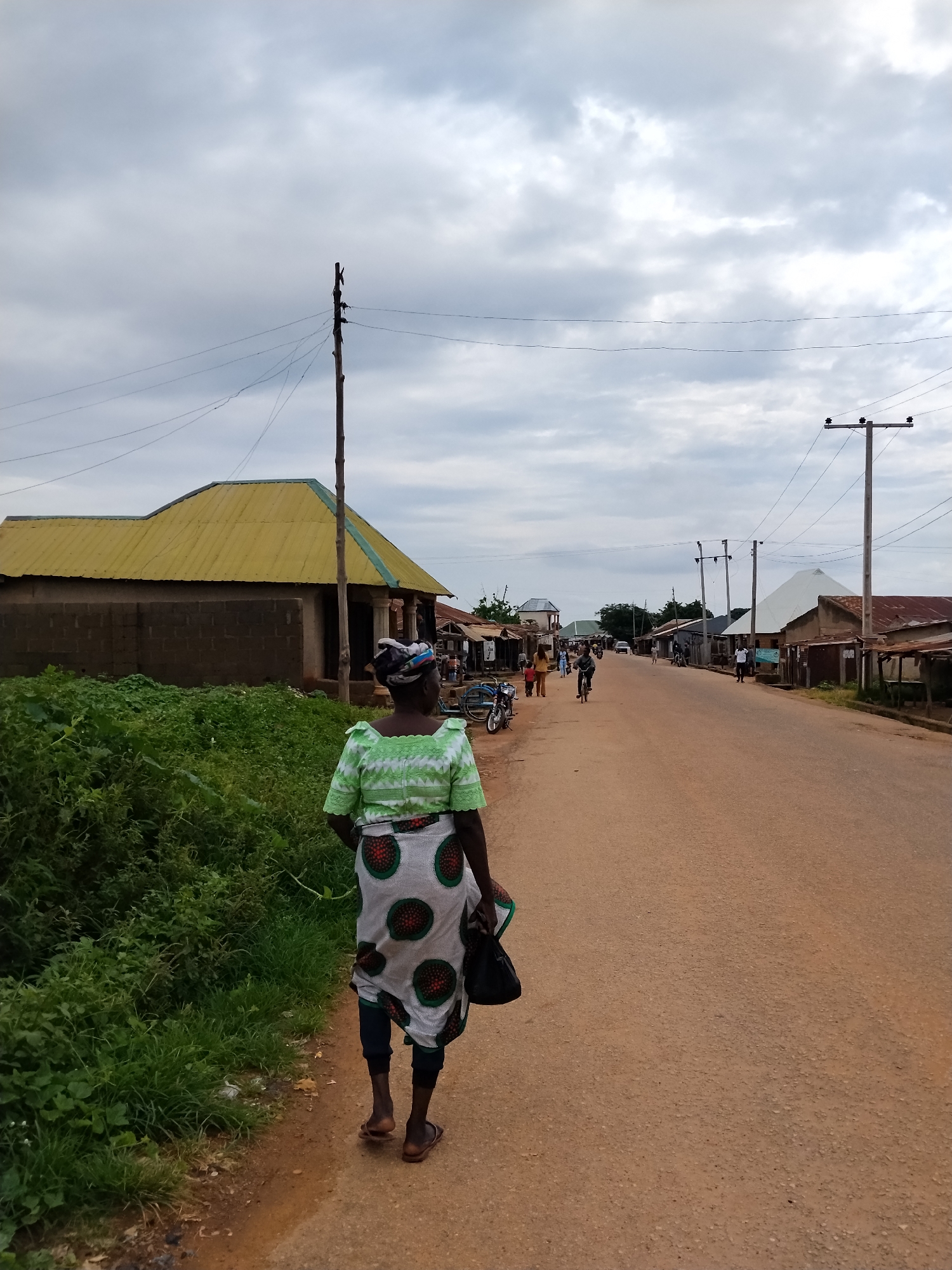 A full shot shows a woman walking away from the camera down a dirt road in Mangu Town, Nigeria. The woman is wearing a light green, patterned top and a white, circular-patterned skirt. She has a dark bag in her right hand. The road is lined with buildings, some of which have corrugated metal roofs. Green vegetation is visible on the left side of the road, and power lines crisscross the cloudy sky. Several other people and a bicycle are visible further down the road, as well as two motorcycles parked on the left.