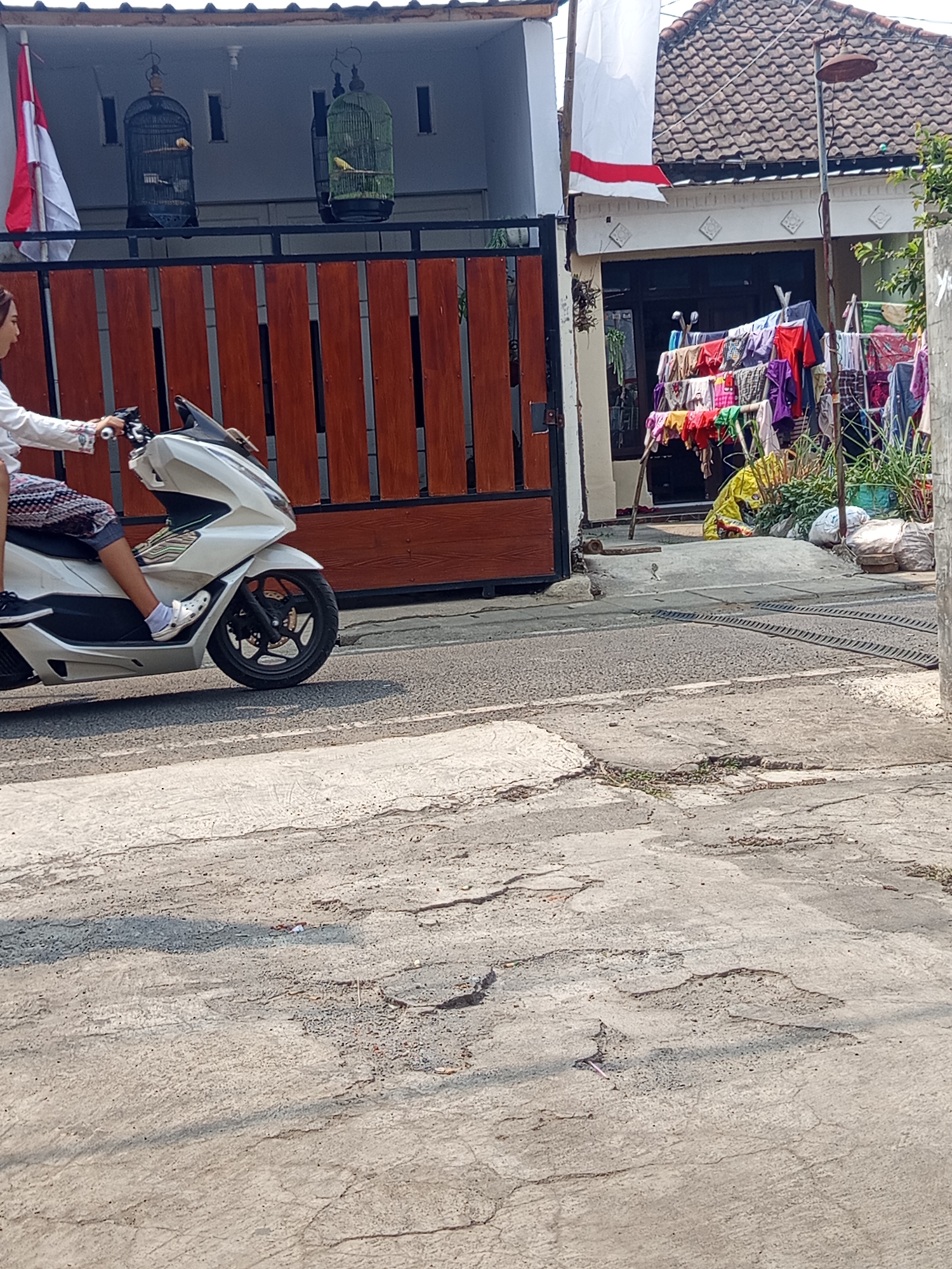 A person is riding a white scooter on a paved road in Malang, Indonesia. The person is wearing a white long-sleeved shirt and patterned shorts, with white Crocs on their feet. To the left of the road, there is a wooden fence and a house with two birdcages hanging from the front. One birdcage contains a light yellow bird, and the other contains a darker bird. A red and white flag is also visible on the left side of the house. To the right of the house, there is a clothesline with various colored clothes hanging on it, as well as some bags and plants. The road appears to be in disrepair with several cracks and potholes.