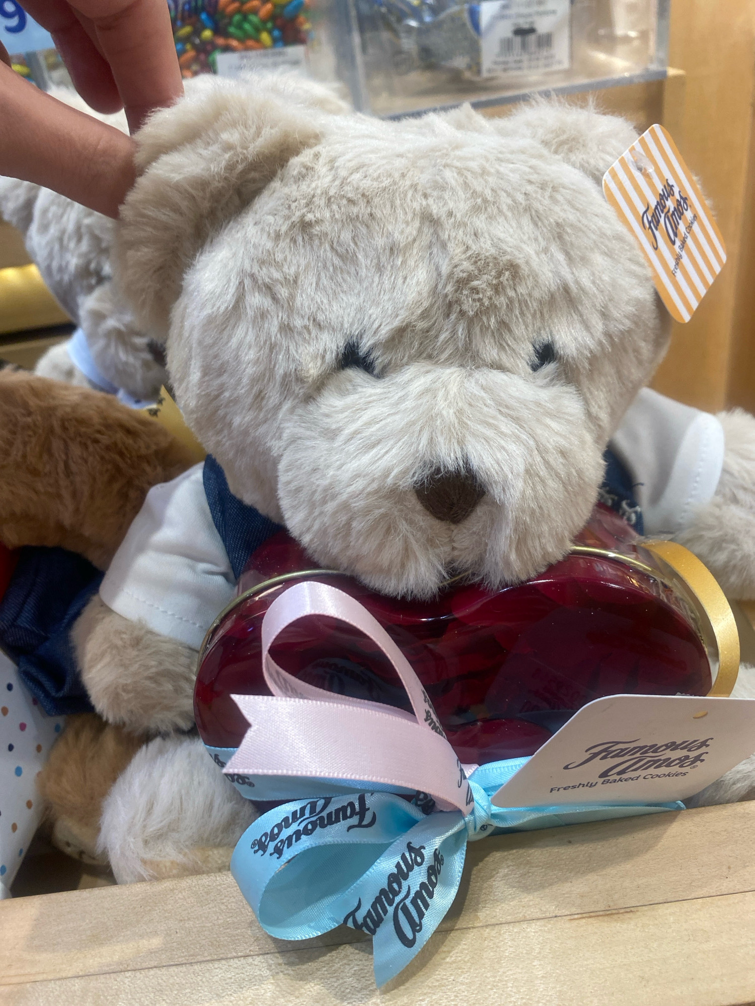 The image shows a close-up of a light beige teddy bear wearing a white shirt and a denim vest. The bear is holding a clear, heart-shaped container filled with red candies or cookies, tied with pink and blue ribbons. A tag attached to the bear's ear reads "Famous Amos Freshly Baked Cookies." Another tag with orange and white stripes is visible on the bear's head. The background is blurred, but appears to be an indoor setting, possibly a store display. No specific location in Kuala Lumpur, Malaysia is identifiable from the image. There are no people clearly visible in the frame, only a hand partially visible at the top left, touching the bear's ear.