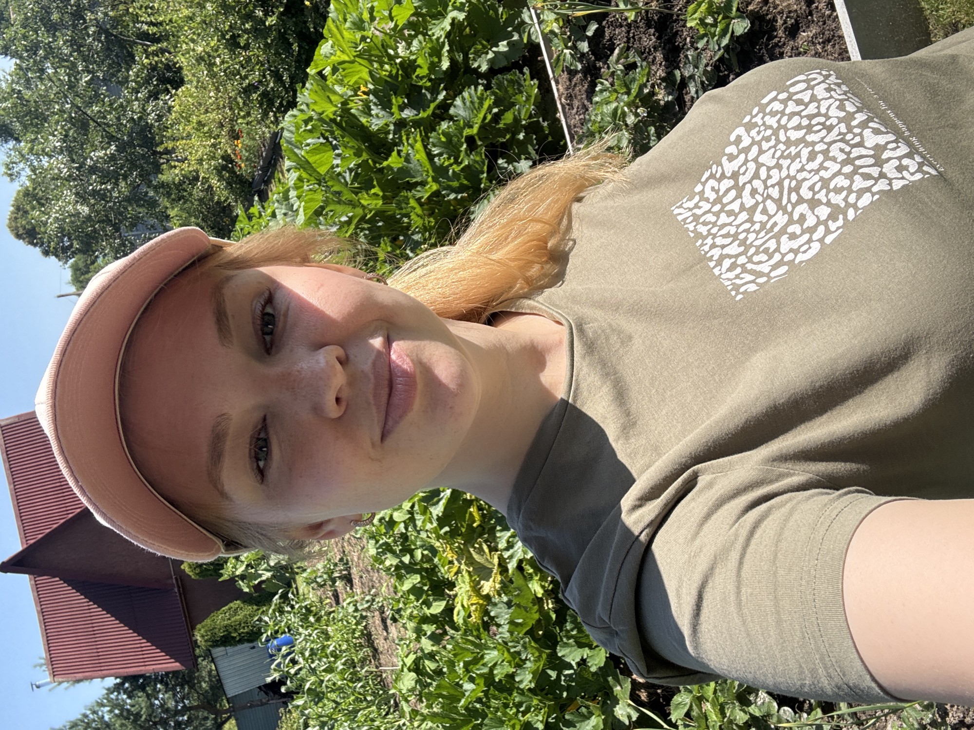 A young Caucasian woman with blonde hair and a pink baseball cap is taking a selfie in an outdoor garden setting. She is wearing a loose-fitting olive-green t-shirt with a white leopard print design on the left chest. The background consists of lush green vegetation, including what appear to be zucchini plants, and a red-roofed building in the distance. The setting is likely a rural area, possibly a dacha or garden plot, in or near the village of Dunilovo, Russia. The lighting suggests it is a sunny day.