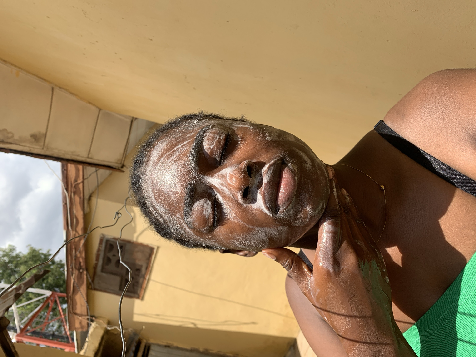 A dark-skinned person with soap on their face is outside against a yellow wall, with their eyes closed and their right hand touching their chin and neck. They are wearing a green and black garment. A thin gold necklace is visible. In the background is a building with visible wires and partly obscured by the building is a cloudy sky and some green trees. The location is Rafin Par, Nigeria.