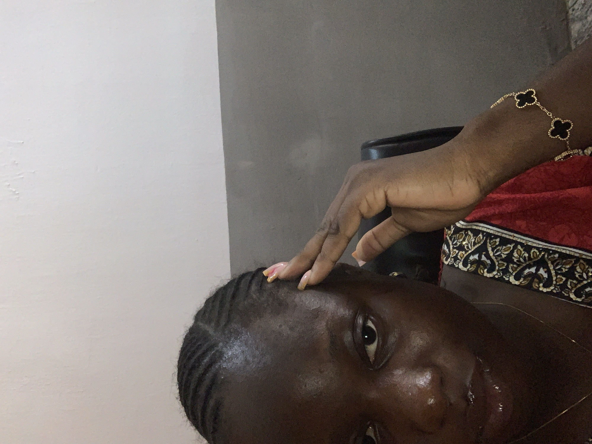 The image is a close-up, waist-up, selfie-style shot of a dark-skinned woman with braided hair. She is looking towards the camera with her right hand touching her forehead. She is wearing a red garment with a patterned trim and a gold bracelet with black clover-shaped charms. Her lips are glossy. The background consists of a light-colored wall and a darker gray wall or partition. The lighting appears to be indoor.