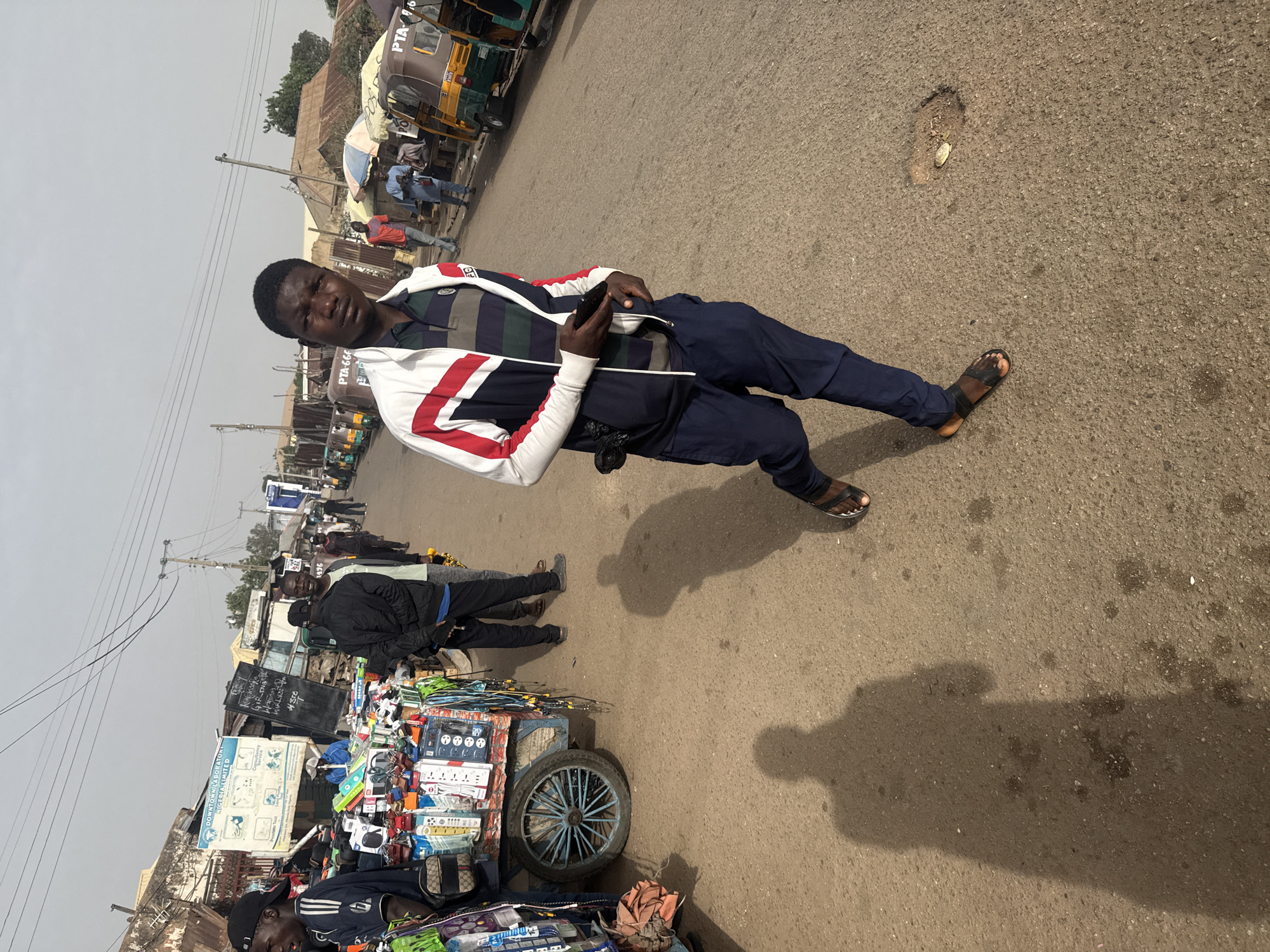 A man stands on a street in Jos, Nigeria. The street is paved and cracked. The man is wearing a jacket with white and red stripes, dark pants, and sandals. He holds a phone. A vendor's cart with various wares sits on the street. Other people and vehicles are visible in the background.