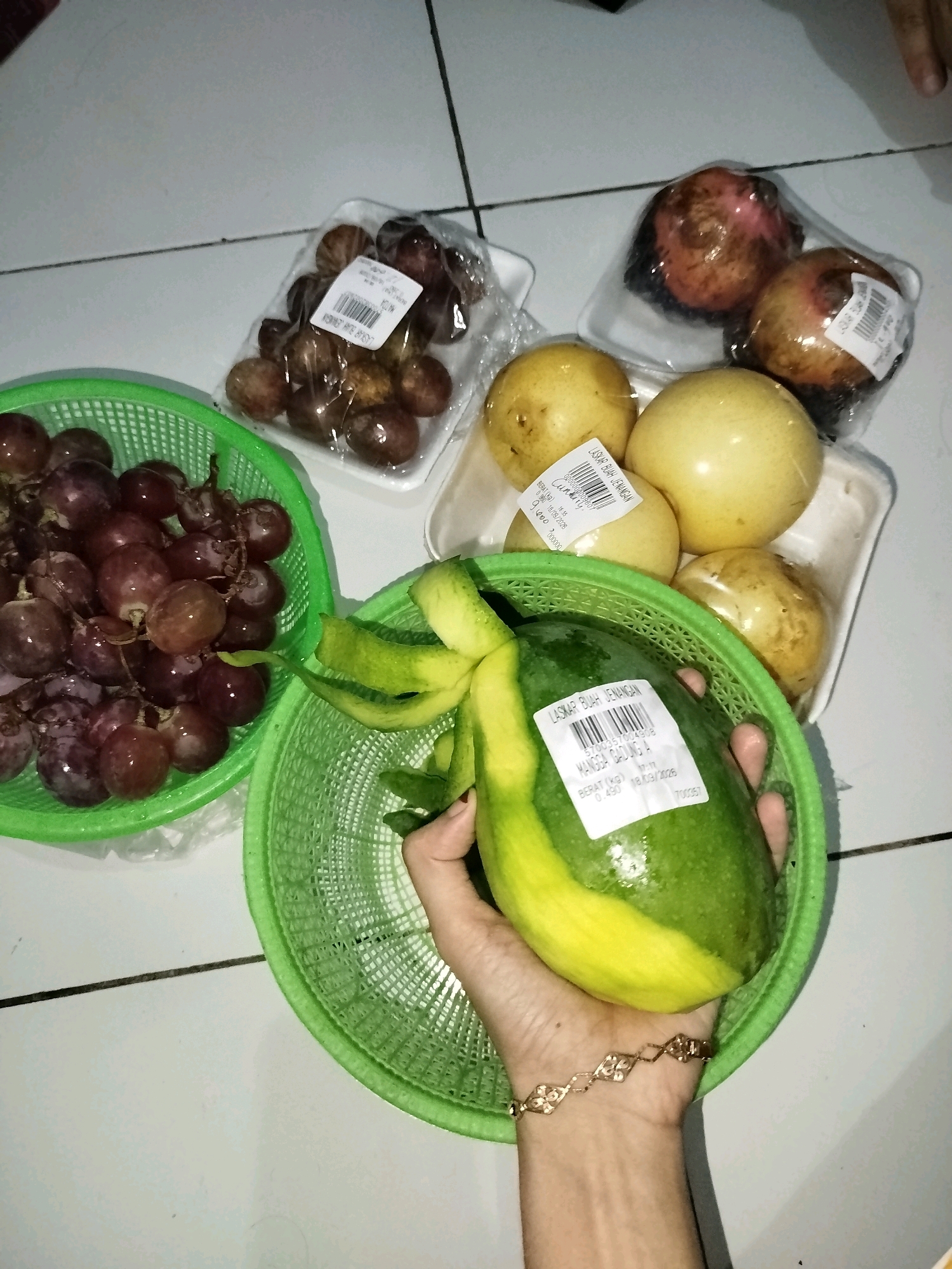 In Babadan, Indonesia, a top-down shot captures an assortment of fruits arranged on a tiled floor. The fruits include a green basket filled with red grapes, another green basket containing a partially peeled avocado with a label, a white tray with four yellow passion fruits, and another white tray with three guavas. The background consists of white and gray tiles, with a dark line intersecting the scene.