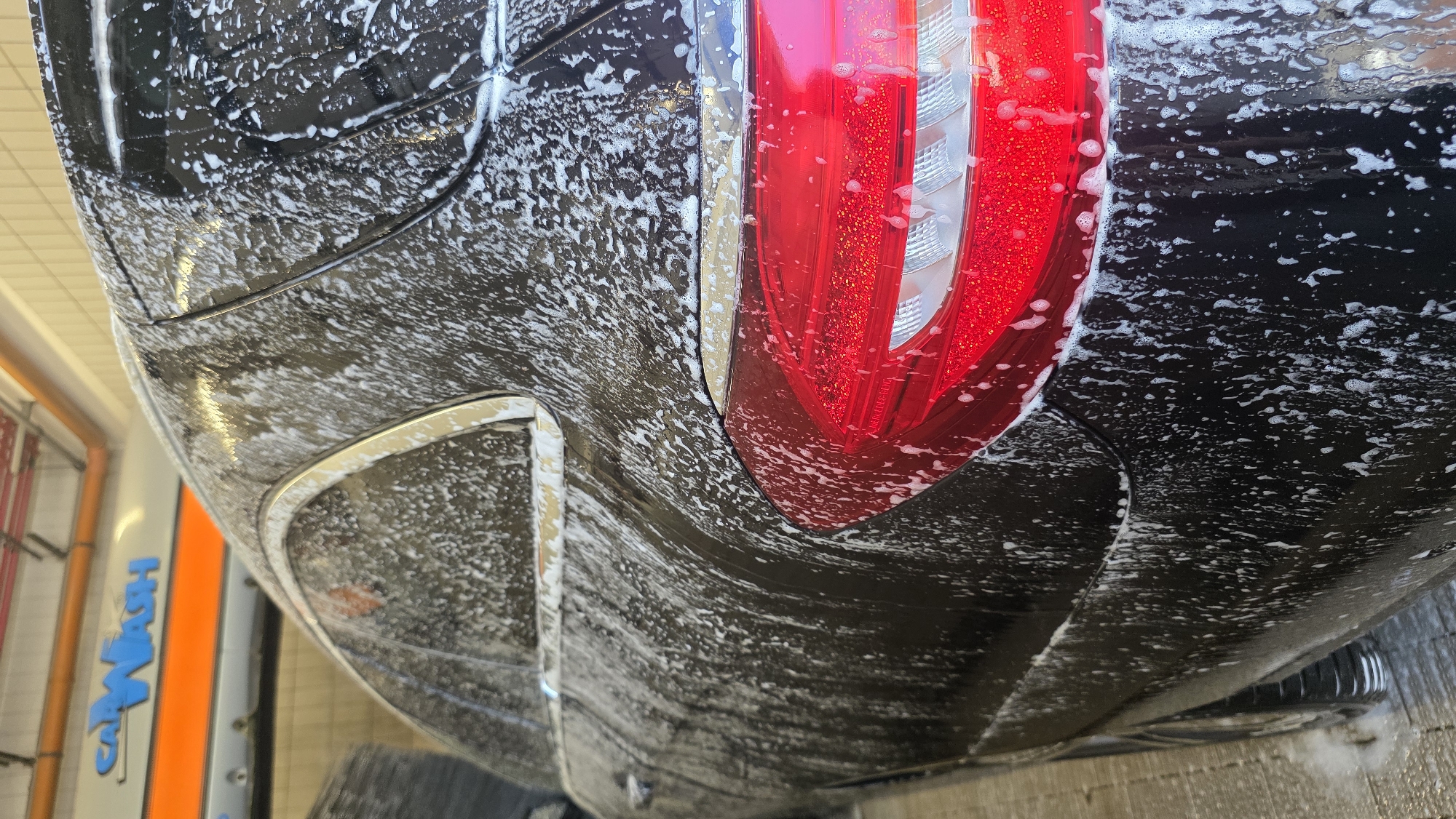 The image shows a close-up, angled view of the rear of a black car covered in white soap suds. The car is wet and appears to be undergoing a wash. The focal point is a red taillight on the right side of the frame, which is partially obscured by the suds. The taillight has a modern, multi-segmented design with visible red and clear/silver elements.
The car's black paintwork is shiny and reflective, showing the distorted shapes of the suds and the surrounding environment. There are distinct curves and body lines visible on the car's rear bumper and quarter panel. The suds are distributed unevenly, creating a textured, foamy appearance across the vehicle.
In the background, to the left of the car, a vertical structure with orange and gray sections is visible. Blue text spelling "CARWASH" is clearly legible on a white panel on this structure. This indicates the setting is likely an automated car wash facility. The lighting suggests it is daytime, possibly indoors or under overcast conditions, as there are no strong shadows. The image does not contain any people or explicit emotional cues, focusing solely on the car and the car washing process.