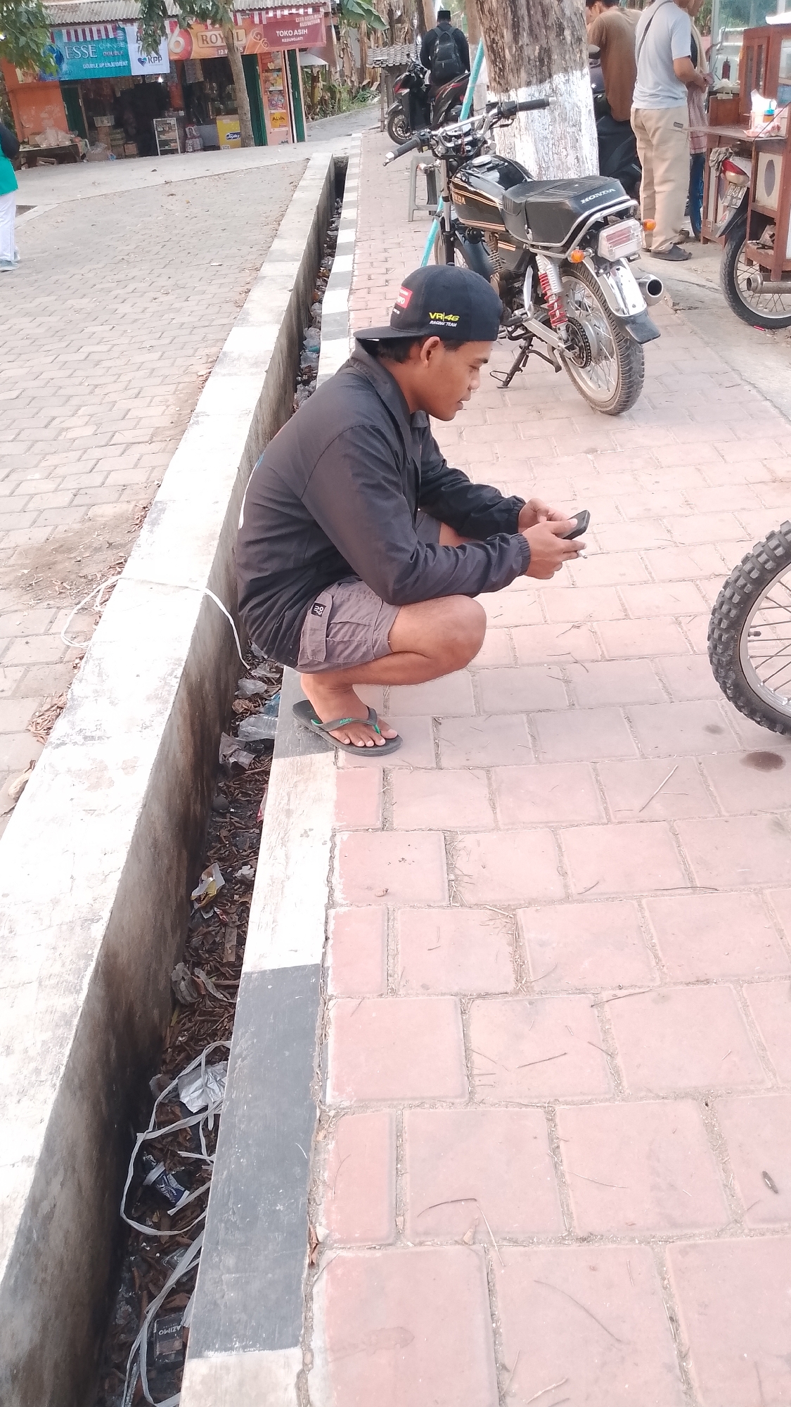 In Karanglangu, Indonesia, a young man wearing a black baseball cap and black jacket crouches on a brick sidewalk, looking at his mobile phone. He is wearing grey shorts and dark flip-flops. Beside him, a black motorcycle is parked. A drainage ditch filled with trash runs alongside the sidewalk. In the background, a building with signs that read "ESSE" and "ROYAL" is visible, with other people and more motorcycles further down the street. The scene appears to be an outdoor public area in a town.
