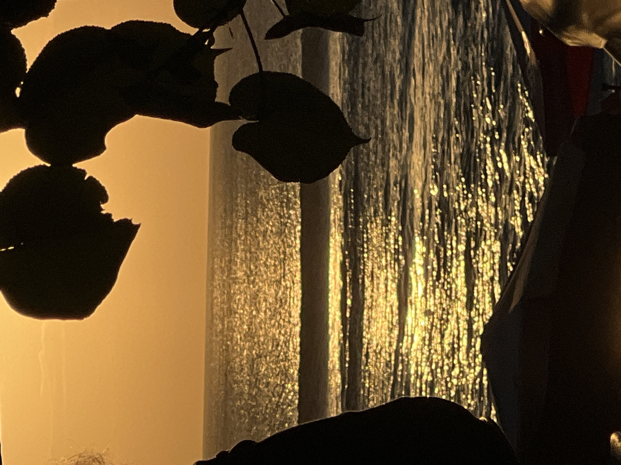 The image depicts a silhouette of a person on the left, standing near the sea. In the foreground, the silhouette of a dark, tent-like structure is visible, with hints of red and yellow fabric. Above the sea, a series of heart-shaped leaves are silhouetted against a vibrant orange sunset sky. The sun's reflection sparkles on the water, creating a shimmering effect. The ocean appears calm with gentle waves breaking on the shore. This scene is likely Kuta Beach in Bali, Indonesia, renowned for its stunning sunsets and beachfront atmosphere.