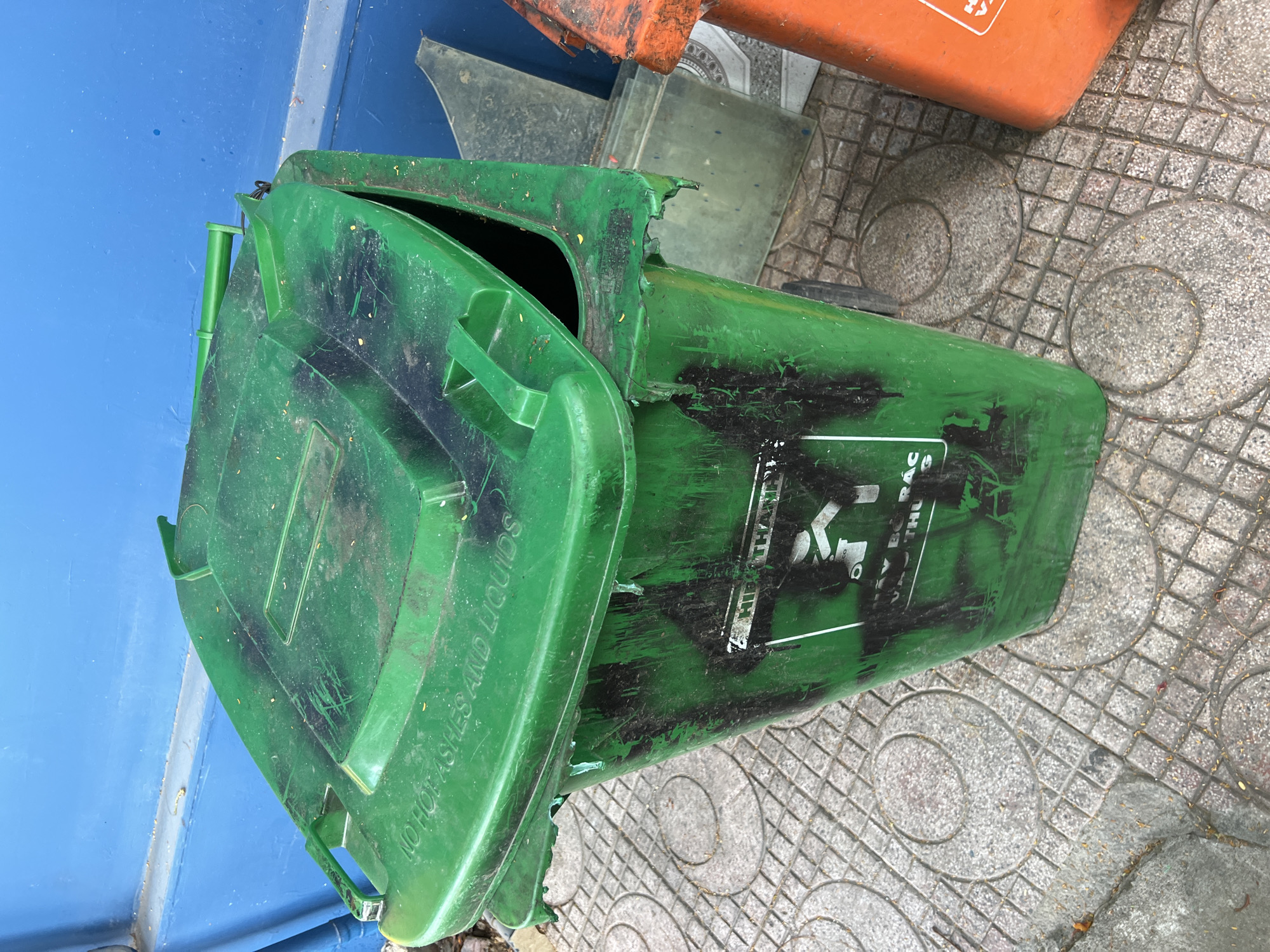 A green, industrial-sized garbage bin with a damaged lid sits on a tiled sidewalk in Nha Trang, Vietnam. The bin is heavily scuffed and marked with black paint or residue. An orange bin is partially visible to the right of the green bin. The background consists of a blue wall. The bin's lid has the words "NO HOT ASHES AND LIQUIDS" embossed on it.