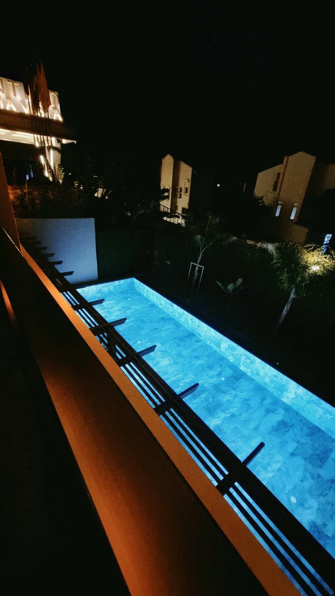 This is a night-time, high-angle, outdoor photograph taken from a balcony or elevated walkway, looking down at a rectangular swimming pool. The pool is illuminated with vibrant blue underwater lights, creating a glowing effect on the patterned tiles at the bottom. The water appears calm and still.
In the foreground, a wooden railing, painted in an orange-brown hue, dominates the left side of the frame and slopes downwards towards the pool. The railing has a textured surface and appears weathered.
Beyond the pool, there is a dark, landscaped area with sparse vegetation and trees. In the background, several modern, multi-story buildings are visible, illuminated with warm yellow lights from their windows, suggesting they are residences or a hotel. One building on the far left has a sign with illuminated lettering, possibly indicating a business or establishment. The sky is completely dark, indicating it is nighttime.
The photograph appears to be taken in the city of Serik, Turkey, given the context of the provided information. The overall scene depicts a tranquil and inviting ambiance, characteristic of a resort or residential area at night. There are no people or discernible animals present in the image.