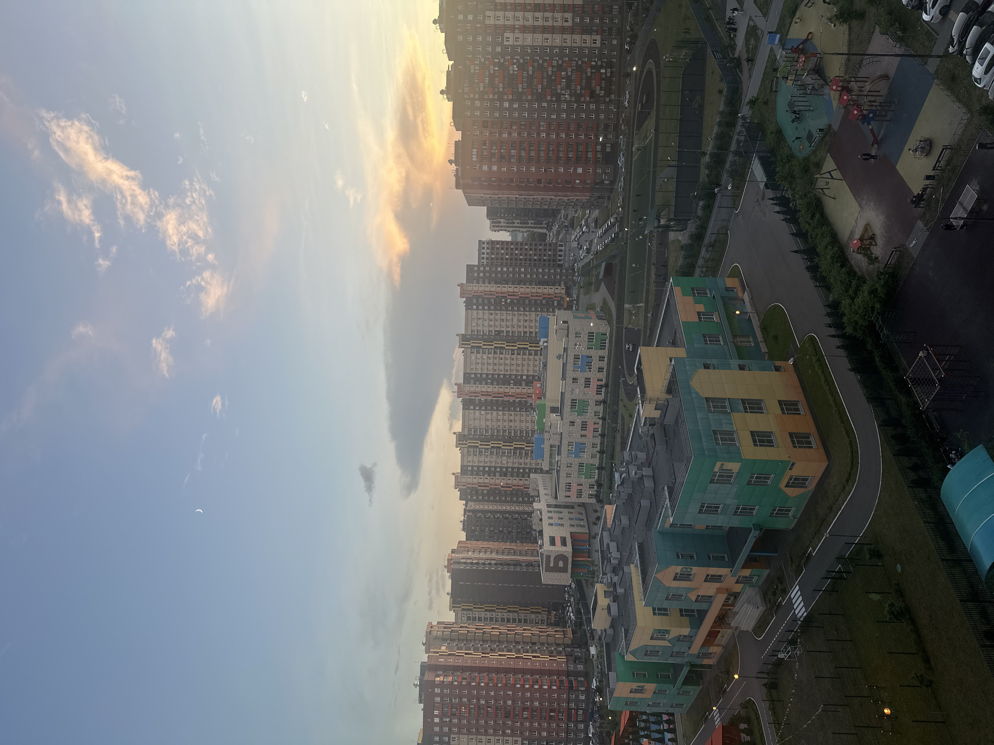 The image captures a twilight scene in the city of Mirny, Russia, featuring a residential area with modern high-rise apartment buildings and a kindergarten. The sky is a gradient of pale blue and orange, with a crescent moon visible and wispy clouds tinged with sunset colors. In the foreground, a brightly colored kindergarten building with green, yellow, and orange facades is prominent, with play areas and sports fields adjacent to it. Several high-rise apartment buildings, constructed with a mix of red, beige, and grey facades, dominate the background. Cars are parked in the streets and parking lots. A few people are visible in the outdoor spaces, with some appearing to be children in the playground and adults walking. The overall impression is of a newly developed urban environment.