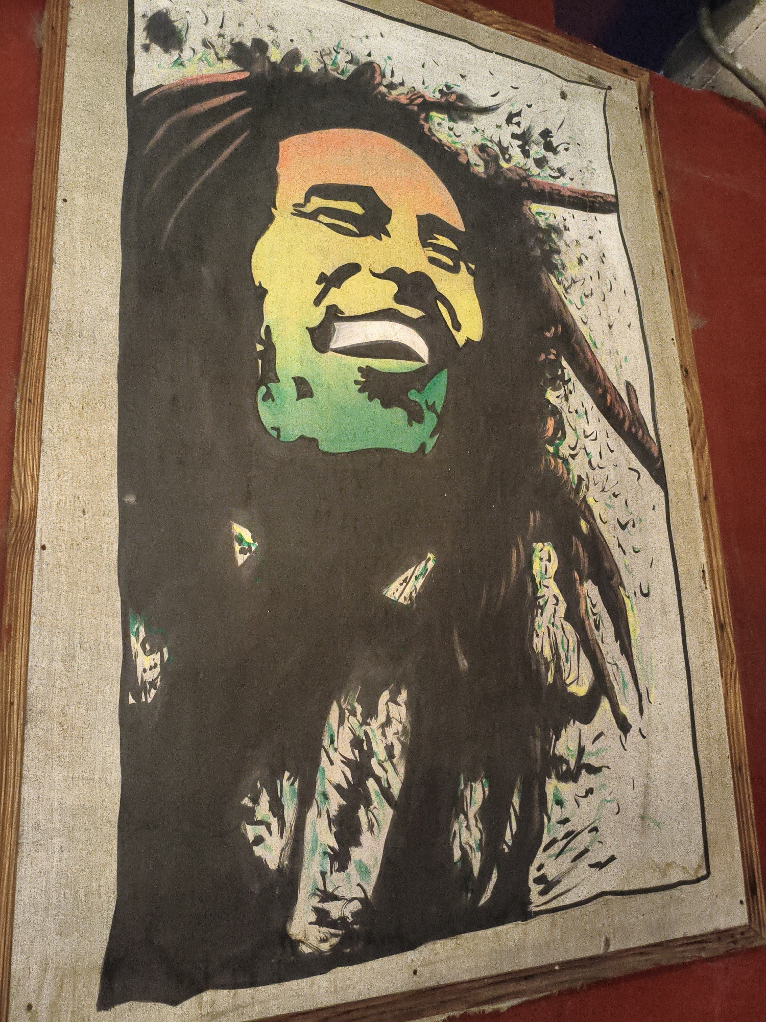 A framed artwork of Bob Marley is displayed on a red wall in Vashisht, India. The artwork is a stencil-like portrait with a black background, featuring Marley's face in shades of yellow, orange, and green. The canvas appears worn and slightly damaged in places. The frame is made of wood and is unfinished. The red wall suggests an indoor setting, possibly a shop or a home.