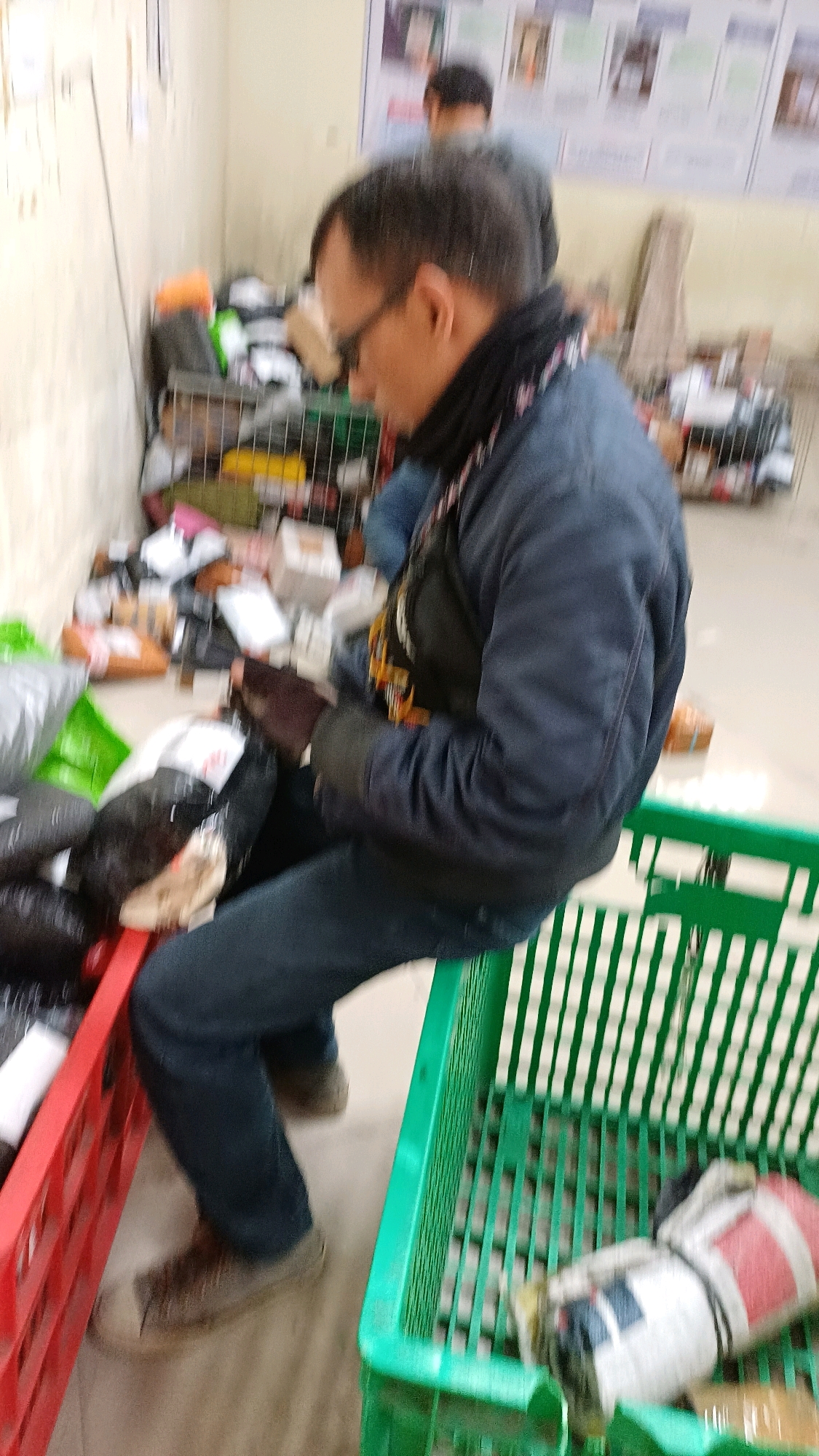 In Sosoran, Indonesia, a person wearing glasses, a dark jacket, and dark pants is sitting on the edge of a red plastic crate, sorting through packages. The person's right hand is gloved and is holding a package. The person is surrounded by piles of packages and two plastic crates, one red and one green. The green crate contains several items, including a cylindrical object wrapped in white and red material. In the background, another person is visible, and there are posters on the wall. The environment appears to be an indoor storage or sorting area.