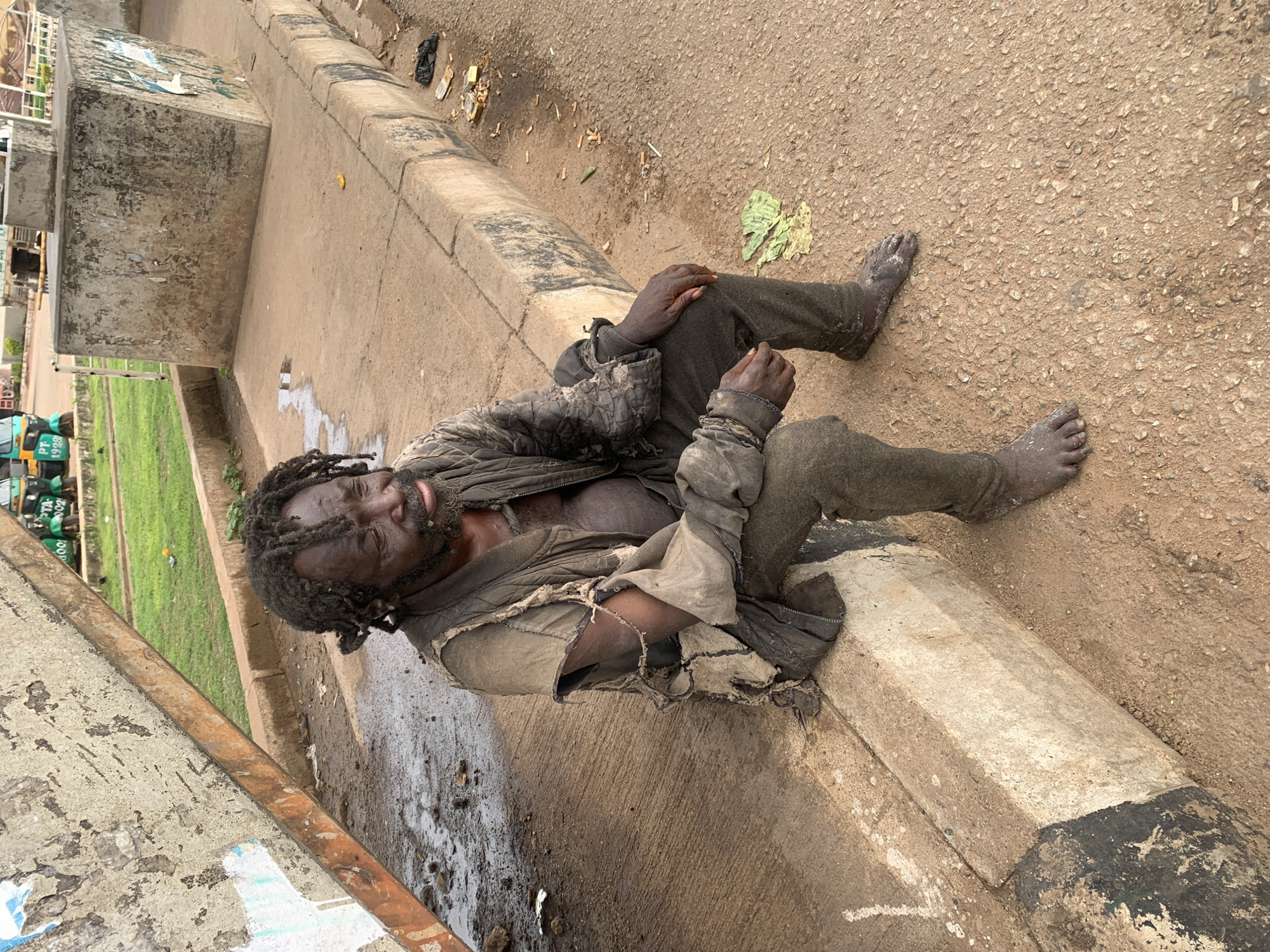 An adult male is observed sitting on a concrete curb in Gura Top, Nigeria, in an urban or semi-urban setting. The individual, appearing to be of Black ethnicity with matted dark hair styled into dreadlocks, exhibits signs of significant distress and neglect. His eyes are closed or heavily lidded, and he possesses a matted beard.
He is clad in multiple layers of heavily soiled, tattered, and torn garments. The outermost layer is a dark, possibly quilted, jacket with significant rips, exposing portions of his chest. Beneath this, a lighter-colored, equally torn shirt is visible, particularly around the shoulders and arms. His trousers are dark, soiled, and appear worn. The man is barefoot, with his feet and hands appearing calloused and heavily caked with dirt. He is seated with his hands resting on his knees, his posture suggesting weariness or introspection.
The immediate environment consists of a concrete sidewalk with muddy patches and debris, separated from a brown, granular road surface by a concrete curb with remnants of black and white paint. Adjacent to the man on the road, there is an accumulation of litter, including numerous discarded cigarette butts, a black plastic bag, various wrappers, and several large green leaves.
In the mid-ground, a large, weathered concrete block, possibly part of a structure or debris, leans prominently on the sidewalk, displaying peeling material and stains. Further back on the sidewalk, another smaller concrete block is visible. The background features an area of green grass and foliage, and two green and yellow auto-rickshaws (locally known as Keke Napep in Nigeria) are partially visible. One auto-rickshaw bears the license plate 'PF 1929'. A white metal railing or fence is also discernible in the background.