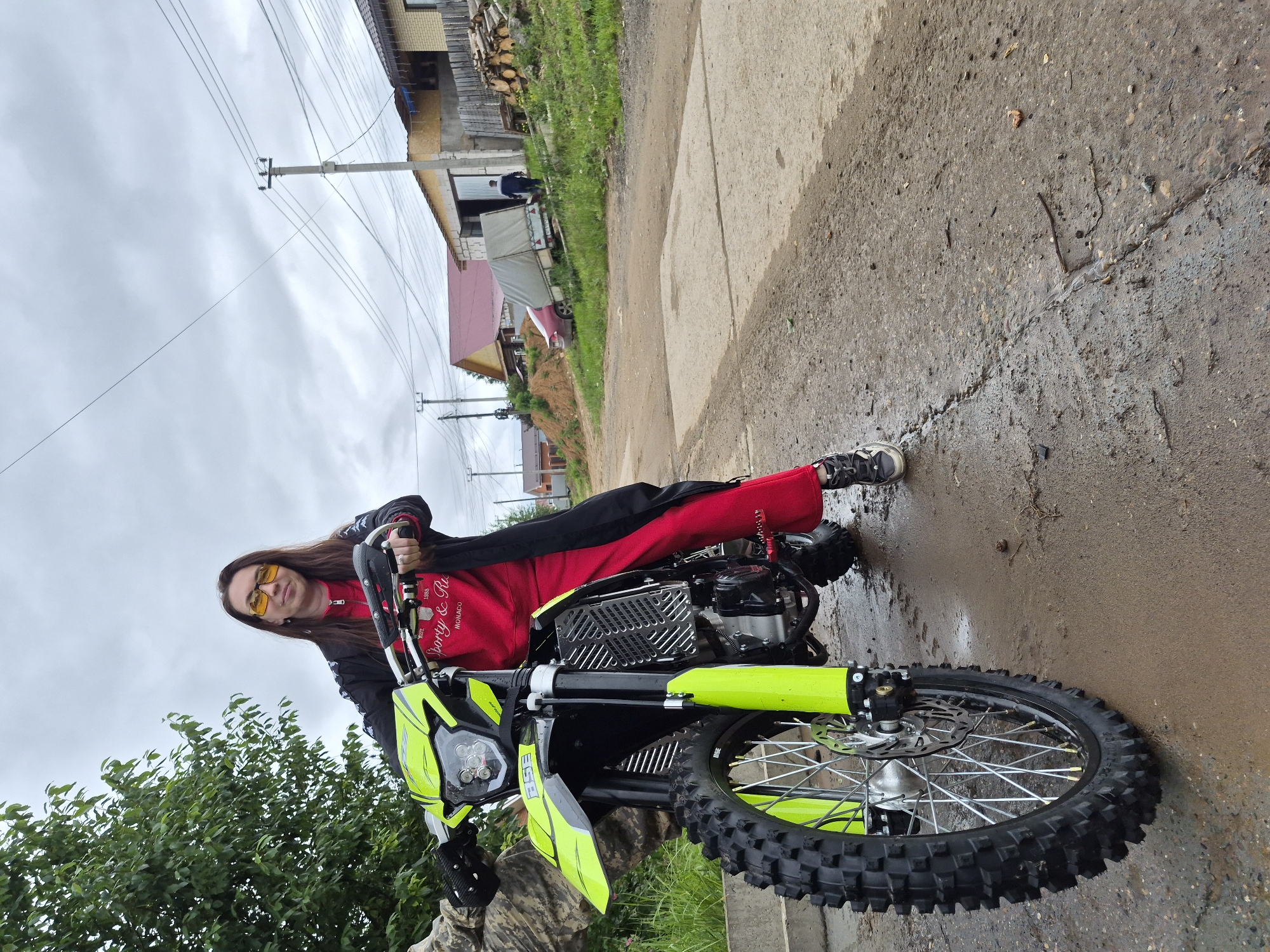 This image captures an outdoor scene in what appears to be a rural or suburban setting in the city of Chaikovsky, Russia, under an overcast sky.
The primary subject is an individual, presumably female, positioned on a fluorescent green and black dirt bike. She is wearing a red long-sleeved top with "EST. 1988 Sporty & Ru MONACO" text, a black jacket, red pants, and yellow-tinted sunglasses. Her long dark hair is visible. Her left foot is on the wet, muddy ground, supporting the motorcycle, while her hands rest on the handlebars. She is looking directly towards the camera. The dirt bike features knobby tires, a front disc brake, a headlamp unit with multiple LEDs, a silver radiator guard, and the brand "BSE" visible on the front fender. Red footpegs are also noticeable.
The ground consists of a wet, unpaved road mixed with sections of cracked concrete or asphalt, exhibiting puddles and muddy areas, indicative of recent rain. Small sticks and debris are scattered on the ground.
In the background to the left, a large tree with dense green foliage is visible. To the right, utility poles with multiple overhead power lines traverse the scene. Several houses or buildings are present, including one with a red roof and another seemingly under construction with exposed framing and a yellow-painted section. A pile of cut wood is stacked near one of these structures. Further down the road, a red car with a covered trailer attached is parked partially on a grassy embankment. A male figure, dressed in dark clothing, is walking away from the camera in the vicinity of the car and trailer. The sky is uniformly grey and overcast.
