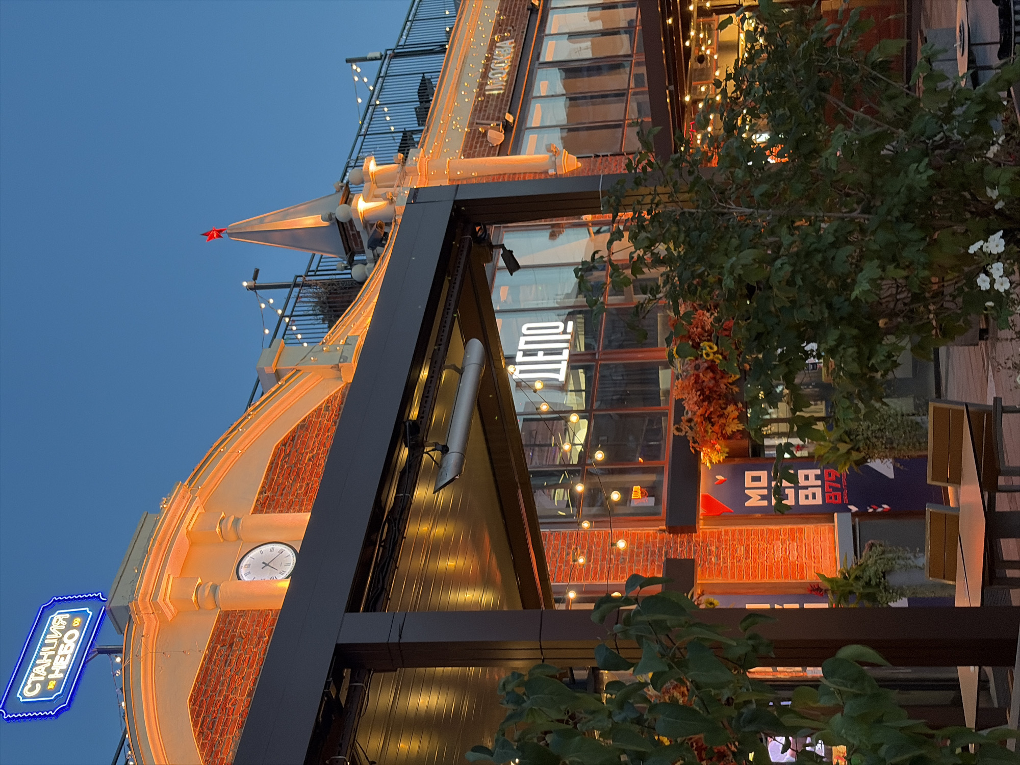 This image depicts the exterior of the "Depo" food mall in Moscow, Russia, during the evening. The building features exposed brickwork, illuminated by warm orange lights, creating a festive atmosphere. A prominent neon sign in blue, reading "СТАНЦИЯ НЕБО" (Sky Station), is visible at the top of the building, along with a classic analog clock.
The foreground is partially obscured by lush green foliage and potted plants, suggesting an outdoor seating area. String lights with round bulbs are visible, casting a soft glow. Through the windows of the building, the interior of the food mall is partially visible, with more lighting and the reflection of the exterior lights.
A red star atop a spire on the building's roof is a distinctive architectural detail. Signs within the building and on banners are visible, including "ДЕПО" (Depo) in large white lettering and "МОСКВА" (Moscow) in Cyrillic. The overall impression is that of a lively and inviting urban entertainment and dining destination. There are no people clearly visible in the image. The sky is a deep blue, indicating twilight.
