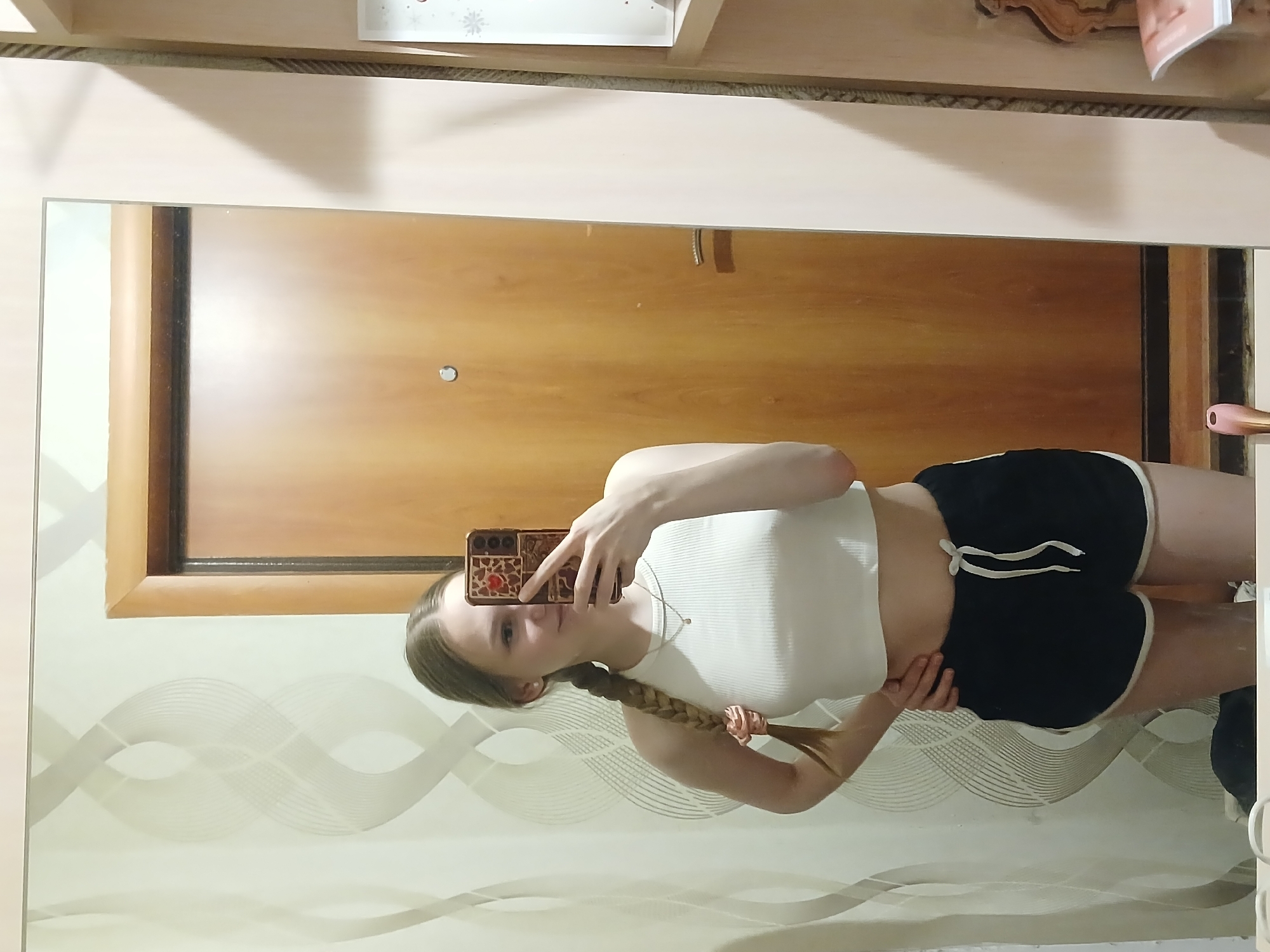 A young woman is taking a selfie in front of a mirror. The mirror reflects a room with a wooden door and a wall with decorative patterns. The woman is wearing a white top and dark shorts with white trim. Her hair is in a braid. The location is presumed to be in a residential setting within the city of Chaikovsky, Russia.