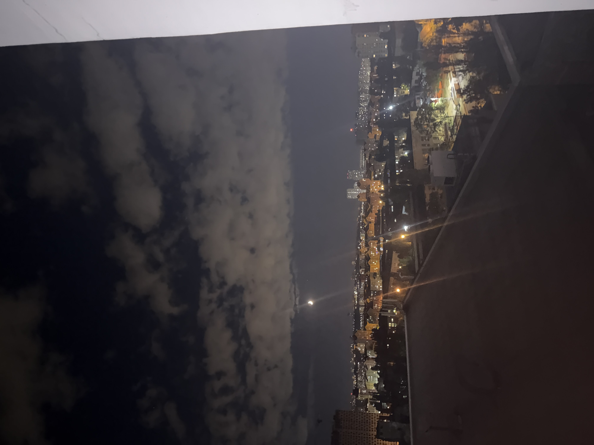 The image depicts a night view of a cityscape, presumably Kyiv, Ukraine, from an elevated position. The sky is dark with scattered clouds illuminated by the moon, which is visible as a crescent shape with a bright halo. Below the sky, the city lights glow, forming a dense urban landscape. Numerous buildings of varying heights and architectural styles are visible, with many windows lit, suggesting activity within. Trees are interspersed throughout the city, their branches silhouetted against the artificial light. In the foreground, a dark, flat rooftop occupies the lower portion of the image, with a white wall on the right side. The overall atmosphere is quiet and serene, characteristic of a nighttime urban scene.