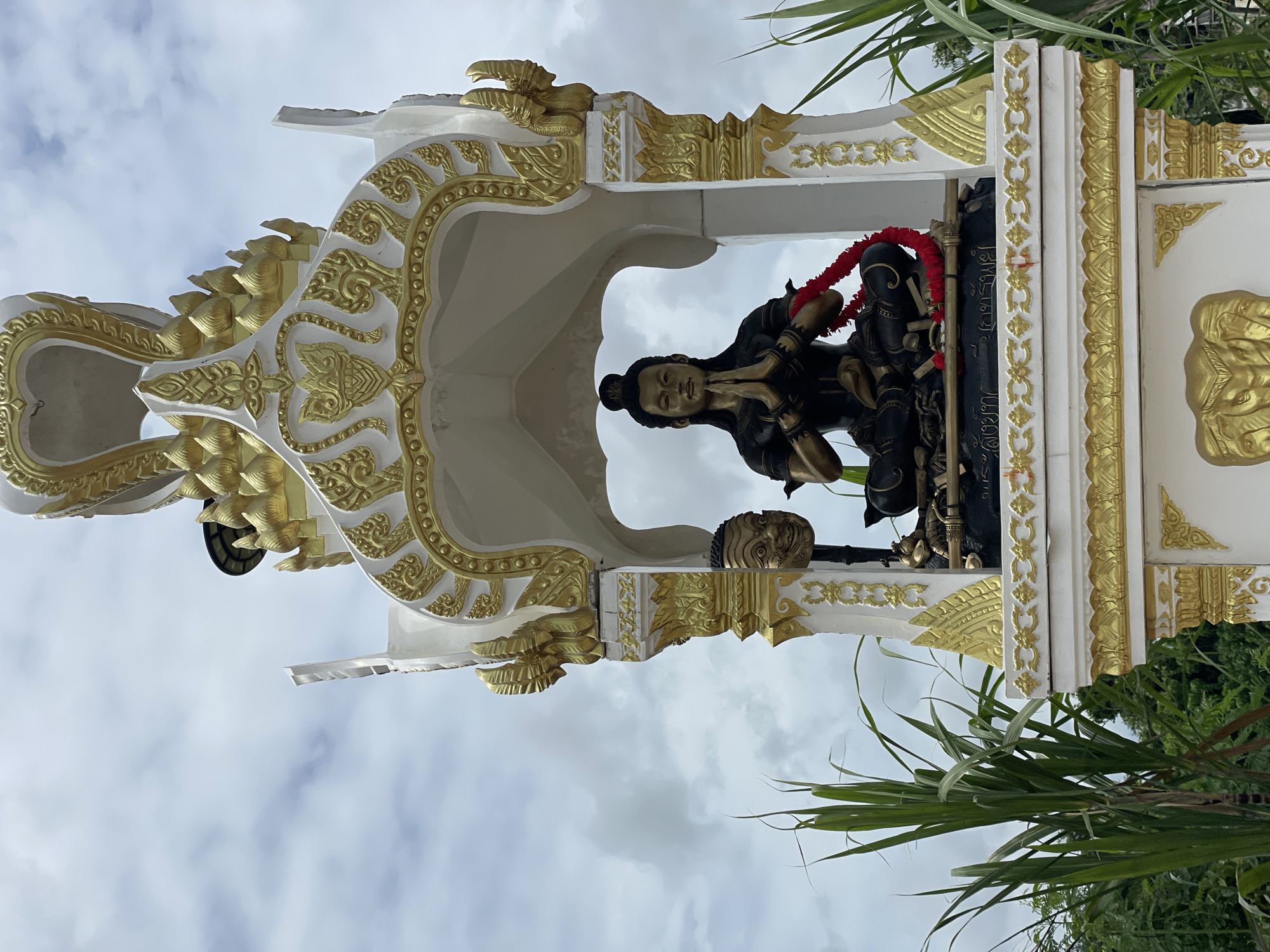 A statue of a seated figure with hands pressed together in prayer is displayed within a white and gold shrine. The figure is dark-colored and adorned with a red garland around its neck. To the left of the main statue, a golden mask with a smiling face is mounted on a pole. The shrine is ornate with decorative gold accents and is situated outdoors, with green vegetation visible on the sides and a cloudy sky above. The shrine appears to be a religious or cultural monument. The location is in Tambon Bo Win, Thailand.