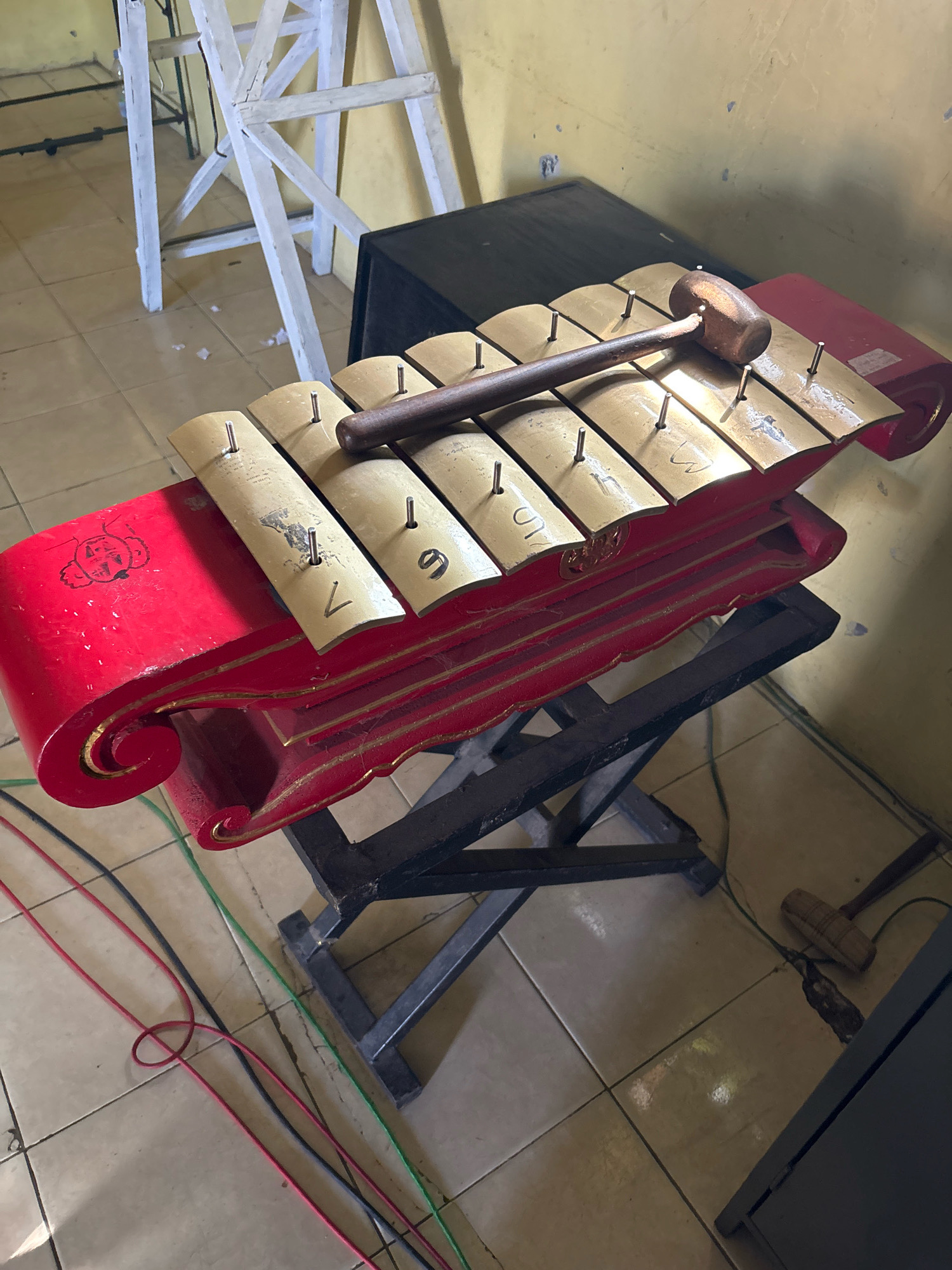 The image shows a gamelan instrument, a type of traditional Indonesian orchestra, placed on a stand. The gamelan has bronze bars of varying sizes, each resting on a red, ornate wooden casing. A wooden mallet, used to strike the bars, lies across the instrument. In the background, a white ladder and a black speaker are visible. The floor is tiled, and several colorful wires are scattered across it. The scene appears to be indoors, possibly in a performance space or practice room, given the presence of musical instruments and sound equipment. The context of Tirtomoyo, Indonesia, suggests this is a cultural setting where gamelan music is performed.