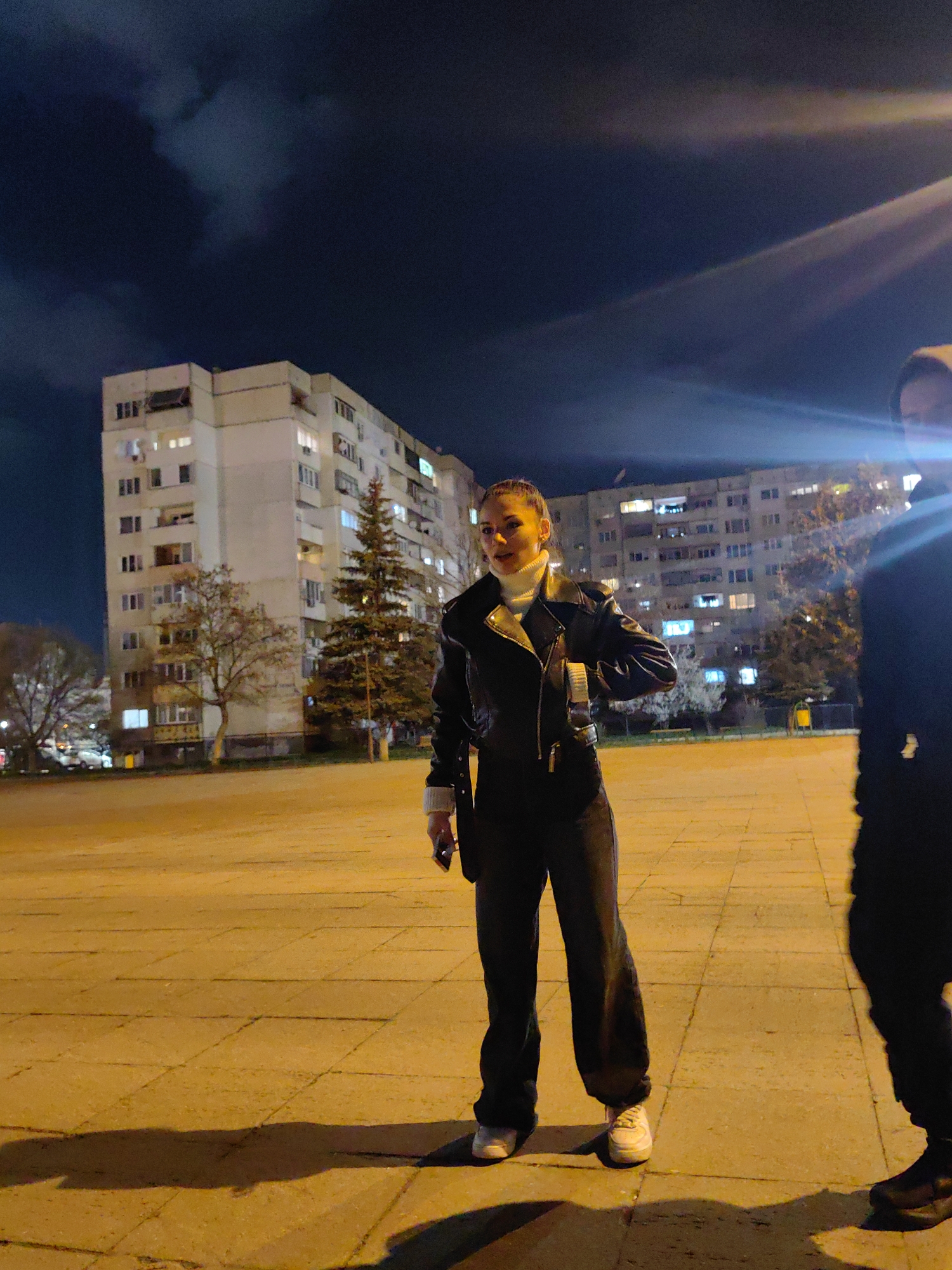 The image captures a nighttime scene in a residential area of Sofia, Bulgaria, characterized by prominent Soviet-era architectural apartment blocks. The sky is dark and cloudy. The ground in the foreground is a paved surface, likely a square or wide sidewalk, illuminated by warm, artificial street lighting, resulting in an orange-yellow hue.
A female individual, appearing to be in her mid-20s to early 30s, stands in the center-left of the frame, facing slightly to her right. Her hair is pulled back, and she is dressed in a white turtleneck sweater, a black leather jacket, dark trousers, and white sneakers. She holds a dark smartphone in her left hand, and her right hand is positioned near her upper torso. Her expression is neutral.
To the far right, a second individual is partially visible, primarily as a dark silhouette, with only their right side, legs, and dark shoes discernible. A bright lens flare from an artificial light source originates from the upper right, partially obscuring this individual.
In the background, two large, multi-story concrete apartment buildings with numerous illuminated windows are visible, confirming human habitation. Between the subjects and the buildings, several trees are present, including a tall, dark evergreen conifer and several deciduous trees. A yellow waste bin is faintly visible near the background buildings. Shadows of the subjects extend towards the lower left of the frame.