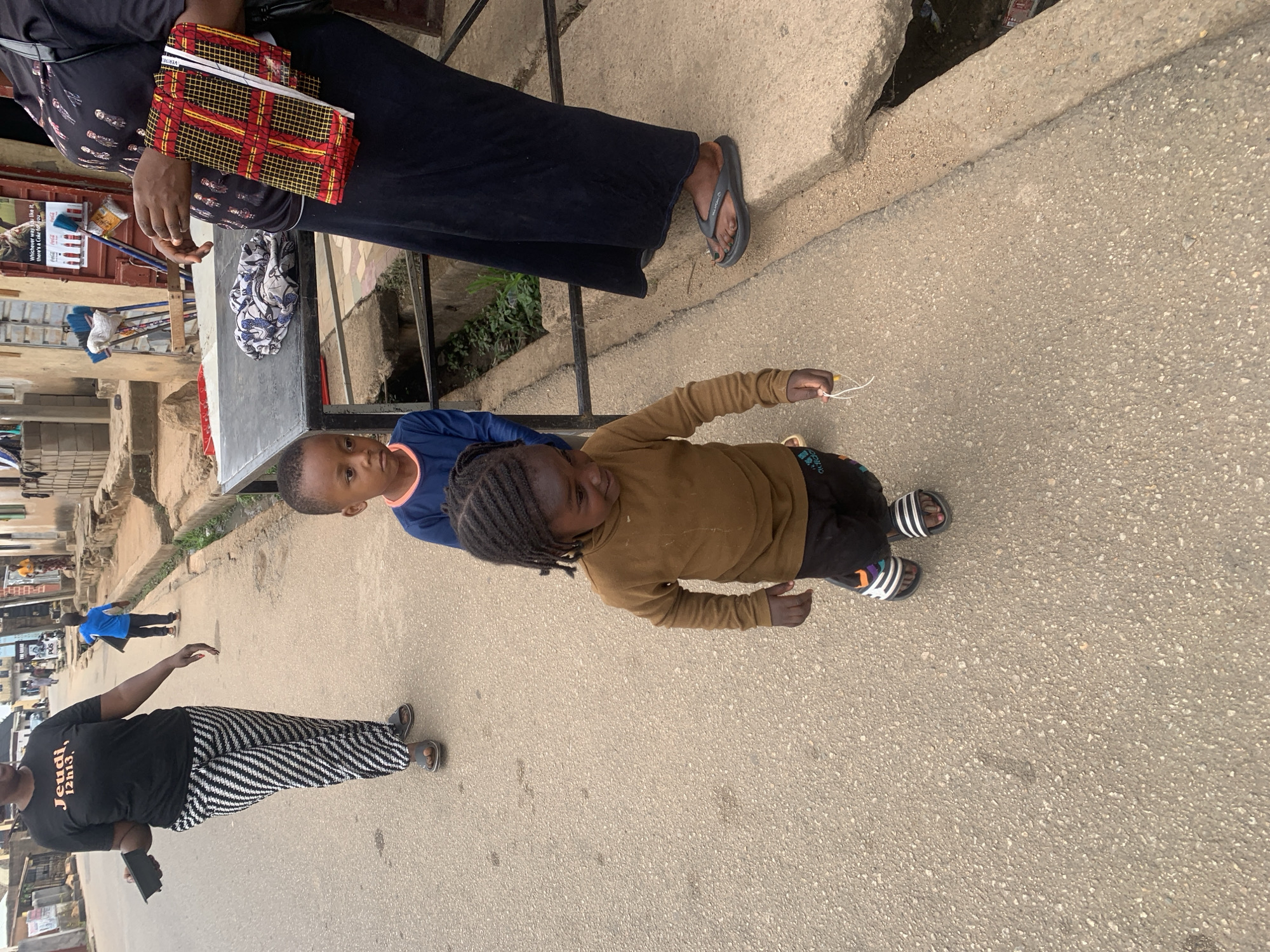 The image depicts an outdoor scene in what appears to be a street in Jos, Nigeria. The street is paved with concrete.
In the foreground, two children are visible. One is wearing a brown long-sleeved shirt and black pants with Adidas sandals and has braided hair. The other child is wearing a blue shirt.
Near them, a person is wearing a black top with text and zebra-striped pants. In the background, another person is walking, wearing a blue shirt and black pants.
In the background are buildings and shops with advertisements, as well as parked vehicles.