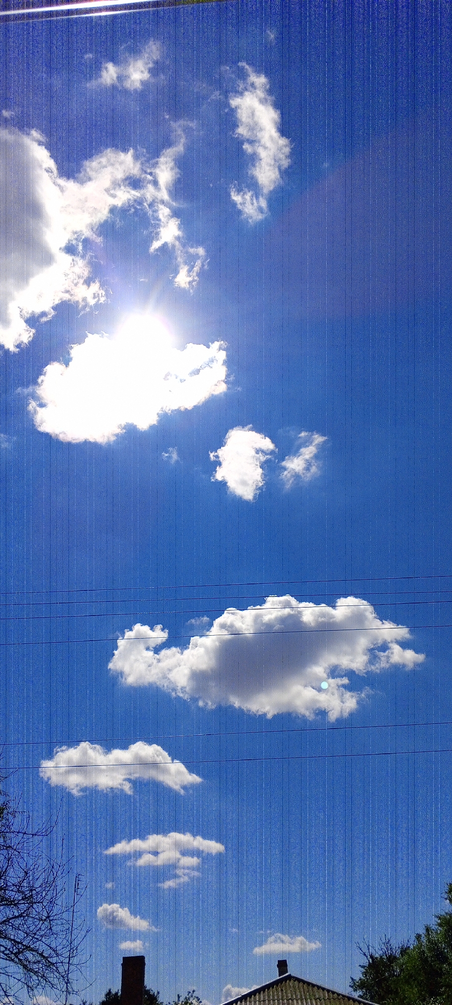The image depicts a clear blue sky with scattered white clouds. The sun is visible, partially obscured by a large cloud, casting bright light. Several thin, dark wires stretch horizontally across the mid-section of the sky. In the lower portion of the image, the tops of several buildings, including a chimney and a sloped roof, are visible, along with some trees. The scene appears to be an outdoor view, likely from a residential area in Novo-Tsimlyanskaya, Russia.