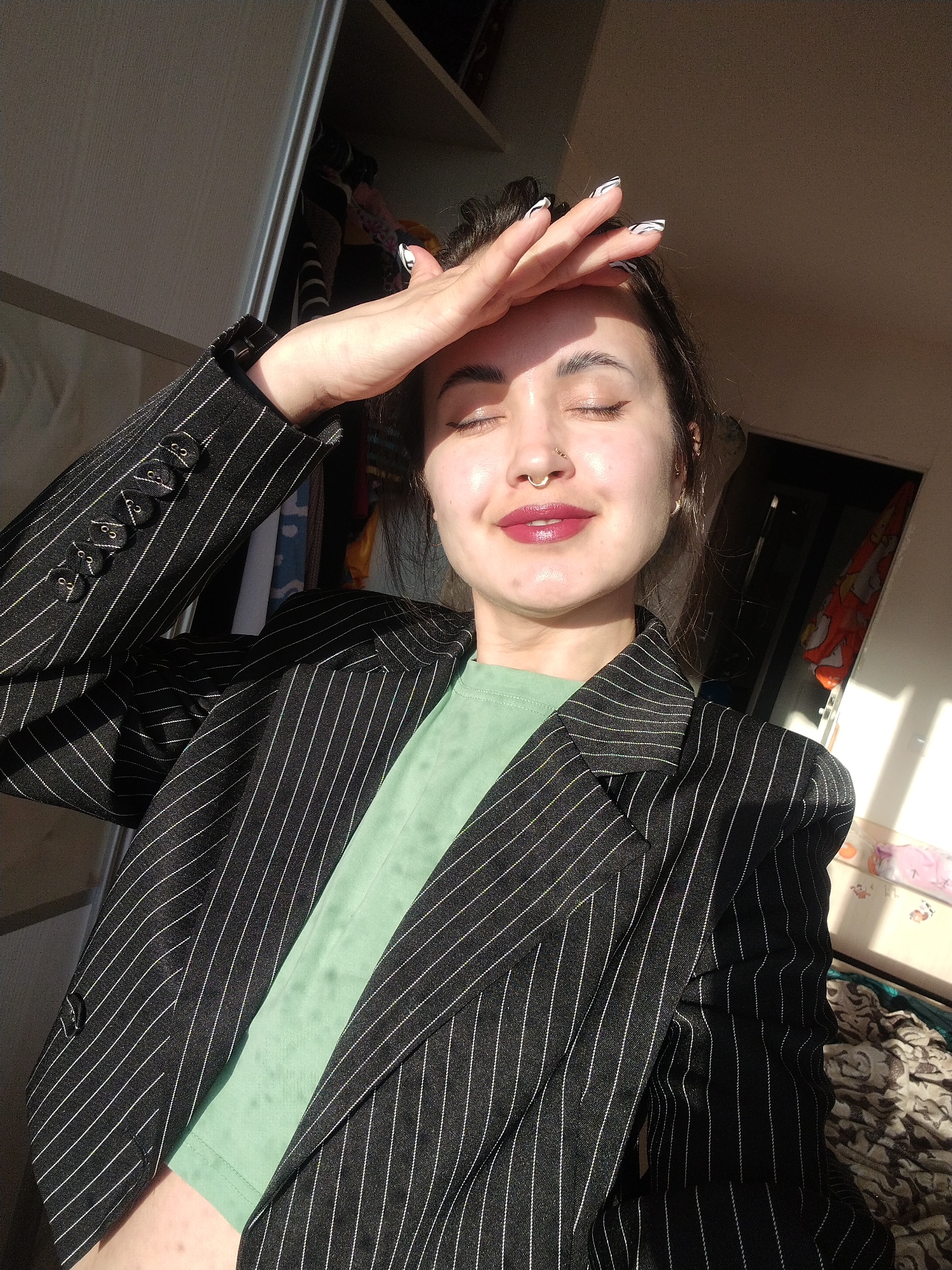 A young adult woman is depicted indoors, illuminated by strong, directional sunlight streaming from the right side of the frame. She is shielding her closed eyes with her right hand, fingers splayed across her forehead, while a faint smile plays on her lips. Her dark hair is pulled back, revealing a golden septum piercing and small earrings. She wears dark pink lipstick and black eyeliner. Her fingernails are adorned with a distinctive black and white zebra-print design.
She is dressed in a black pinstripe blazer with thin white vertical stripes and multiple decorative buttons on the sleeves. Beneath the blazer, she wears a light green crop top. The strong sunlight creates prominent highlights on her skin and clothing, as well as distinct shadows.
The background on the left shows an open closet or wardrobe with various clothes hanging inside. The right side of the background is blurred but appears to be an indoor space, possibly a room with a bed or other furniture covered by fabric. The intense sunlight indicates it is daytime, likely morning or late afternoon. There is no visible text in the image.