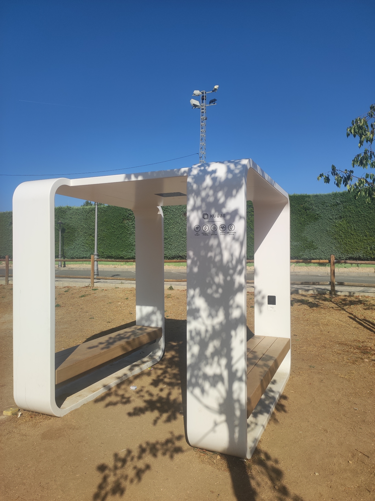 A modern, white, open-air structure with a curved roof and vertical supports is situated in an outdoor area with a sandy ground cover. This structure has two wooden benches integrated into its design. On the right vertical support, a dark rectangular panel is visible. On the left vertical support, there is a white panel with icons and text indicating "KUUBE," and the icons represent Wi-Fi, smartphone charging, and solar-powered ambient sensors. The background features a dense green hedge, a wooden fence, a paved area, and a street with lampposts. In the distance, a tall metal tower with multiple lights stands against a clear blue sky, and a faint contrail is visible. The scene is illuminated by sunlight, casting shadows from the tree's foliage onto the structure and ground. This structure appears to be a public amenity, possibly a charging station or rest area, located in the city of Real Sitio de San Ildefonso, Spain.