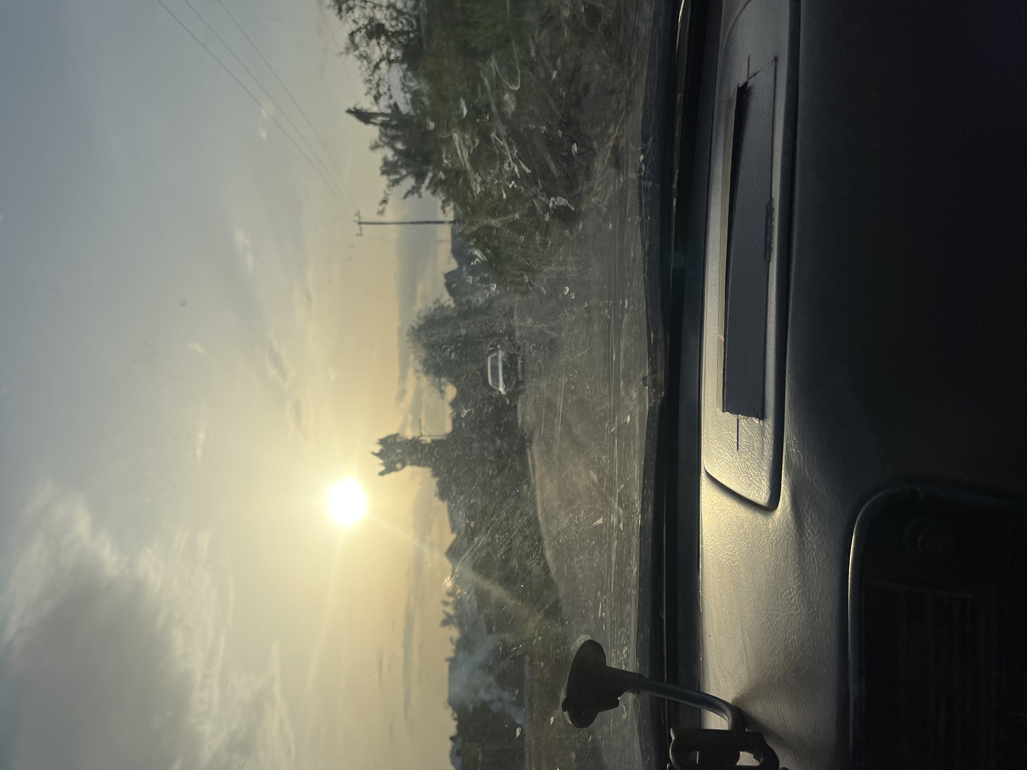 The image presents a perspective from inside a vehicle, looking out through the front windshield towards a road scene at either sunrise or sunset. The sun is a dominant, brilliant white-yellow orb, positioned low in the sky towards the left of the frame, casting a strong glare and contributing to a hazy atmosphere.
The interior of the vehicle is visible on the right side, showing a section of a dark grey dashboard or door panel. A black, suction-cup mounted device, possibly a GPS or phone holder, is attached to the windshield at the lower right. The windshield itself is visibly dirty, covered with numerous streaks, spots, and reflections that scatter the intense sunlight and obscure clarity.
Beyond the dirty glass, a road stretches into the distance, flanked by dense green and brownish vegetation, including trees and bushes. Several utility poles with overhead power lines are visible against the sky, extending across the upper portion of the frame. In the far distance, faint outlines of what appear to be buildings or other structures, and possibly a white vehicle, are discernible through the atmospheric haze and sun glare.
The sky is a mix of bright light around the sun, transitioning to a softer blue-grey with scattered clouds further from the glare. The overall impression is one of a dusty or hazy environment, characteristic of a rural or semi-urban road during a low-sun period. The setting aligns with the provided context of Osogbo, Nigeria. There is no discernible text in the image.