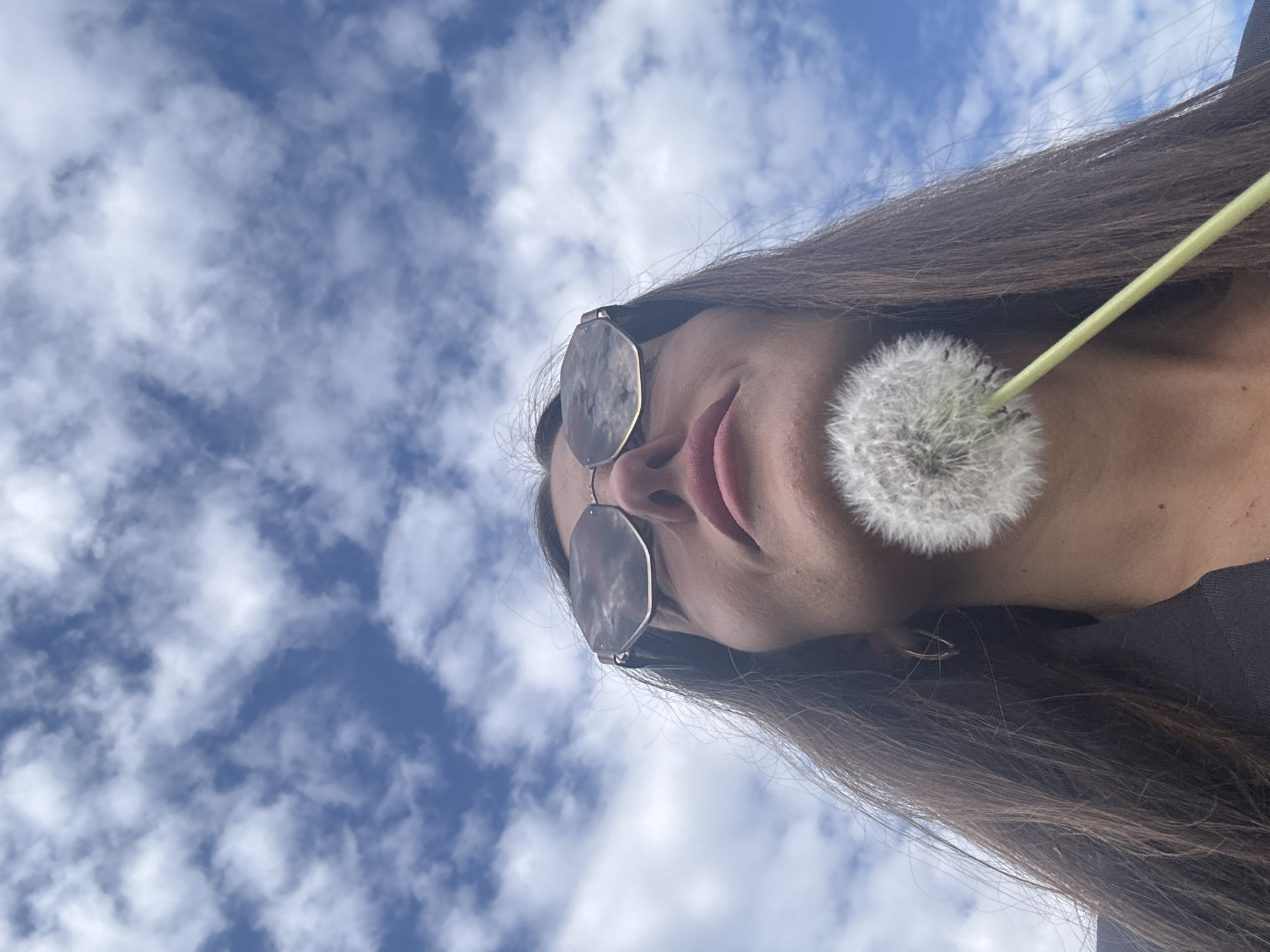 A low-angle, close-up shot captures a person, likely female, looking upwards against a bright blue sky. The view shows the lower half of their face, including the nose, mouth with pinkish lips, and chin. They are wearing hexagonal-shaped sunglasses with dark, possibly reflective lenses and a light-colored metal frame. Long, dark brown hair frames their face and extends downwards.
A fully mature dandelion seed head, often called a dandelion clock, is held close to the person's mouth, with its thin green stem extending upwards past their right ear. The background consists of a clear blue sky dotted with scattered white, fluffy cumulus clouds, indicating an outdoor setting on a bright, sunny day. The image suggests a moment of interaction with nature, possibly the act of blowing on the dandelion for a wish. No visible text or specific geographical landmarks are present in the image, though the context indicates the location is Chaikovsky, Russia. The mood conveyed is serene and engaged with the natural surroundings.