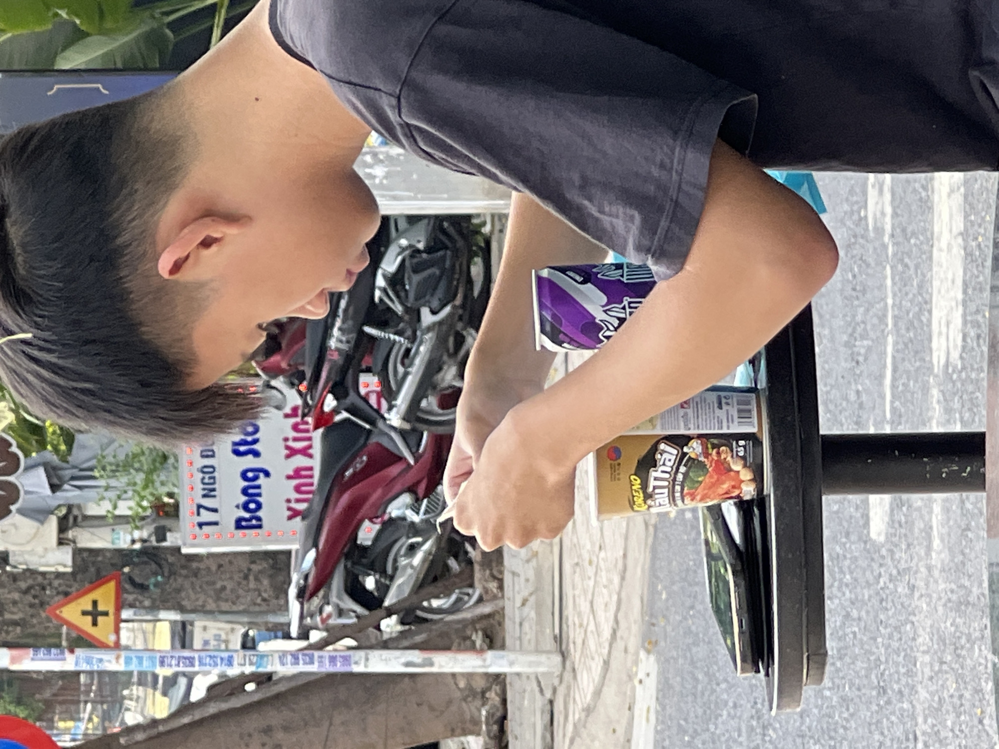 The image captures a young male, seen from the right side of his profile, seated at a small, round, black table outdoors. He has short, dark hair styled with an undercut. He is wearing a dark gray t-shirt. His hands are occupied with a small white object, possibly a piece of paper or food, which he is holding and manipulating. On the table, in front of him, are several items: a can of a purple beverage with a distinctive wave-like design, a cylindrical container of what appears to be snacks with a label reading "KORENO Lẩu Thái" and some Vietnamese text below, and a black smartphone or tablet positioned horizontally. The background shows a street scene in Nha Trang, Vietnam, with motorcycles parked and a traffic sign visible on a pole. The atmosphere suggests an urban setting, likely a roadside cafe or food stall.