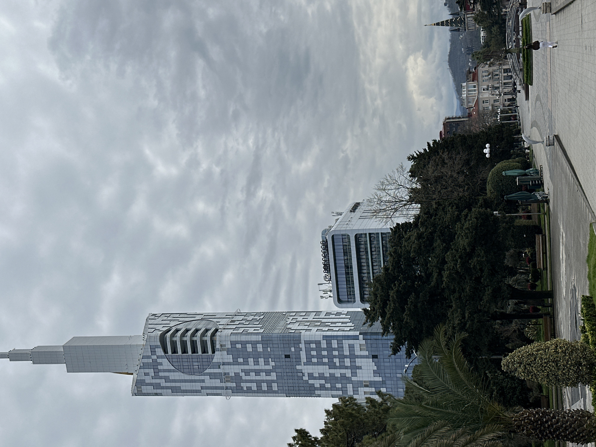 The image depicts a cloudy day in Batumi, Georgia. Prominently featured are two modern buildings: the Alphabet Tower, characterized by its unique architectural design with silver and white geometric patterns and a distinct oval window-like structure near its upper portion, and another high-rise building with "Alliance" displayed at the top. Lush green trees, including palm trees, add a touch of nature to the urban setting. A pedestrian walkway, adorned with dolphin statues and green hedges, stretches into the distance. The backdrop includes other buildings and a glimpse of mountains, all under a gray sky. A person dressed in white is walking on the pathway in the distance.