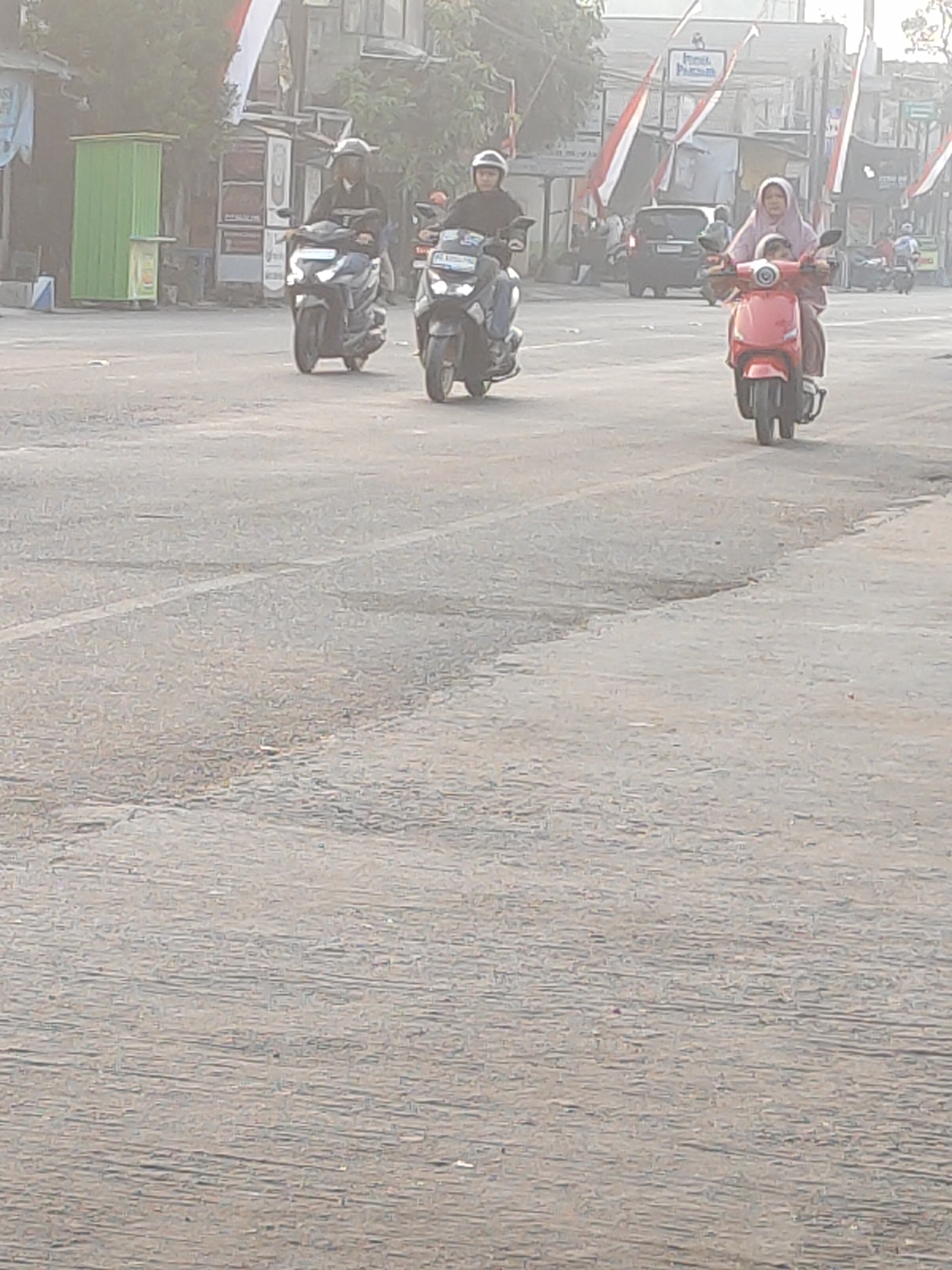The image depicts a street scene in Kanigoro, Indonesia, characterized by a slightly hazy atmosphere, suggesting either early morning or a dusty environment. Three motorcycles are visible on a paved road. In the foreground and slightly to the left, a dark-colored motorcycle with a rider wearing a helmet and dark clothing is moving away from the viewer. To its right and slightly further back, another dark motorcycle with a rider in a helmet and a lighter colored jacket is also moving away. In the mid-ground and to the right, a bright red scooter carries two individuals. The person in front is wearing a light purple headscarf and jacket, and the person behind is also dressed in similar light purple attire. Both are facing forward. In the background, several buildings and shops line the street, some adorned with red and white flags, indicating a festive or commemorative occasion. A green structure, possibly a kiosk or small shop, stands on the left side of the road. The road surface shows some signs of wear and tear, with visible cracks and uneven patches. The overall scene conveys a sense of everyday activity in a bustling Indonesian town.