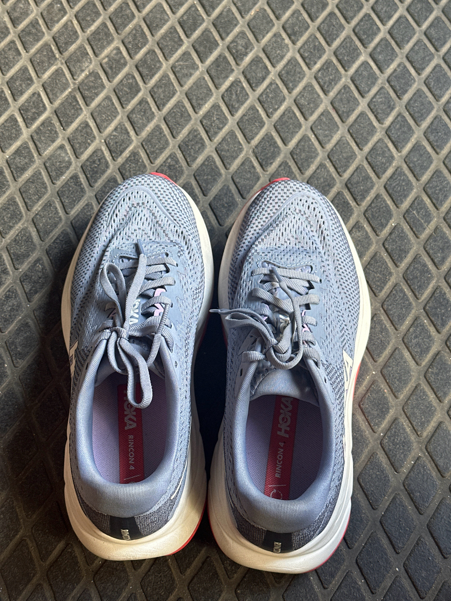 Two light blue running shoes, identified by the "HOKA RINCON 4" branding on the insoles, are placed side-by-side on a dark gray, textured mat with a diamond pattern. The shoes are positioned with the toes pointing upwards. The mat's surface is dry and shows some light wear. There are no people visible in the image. The scene appears to be indoors or in a shaded outdoor area, as there are no strong shadows or direct sunlight. The location is not specified to be in Lalitpur, Nepal, as there are no visual cues to suggest this.