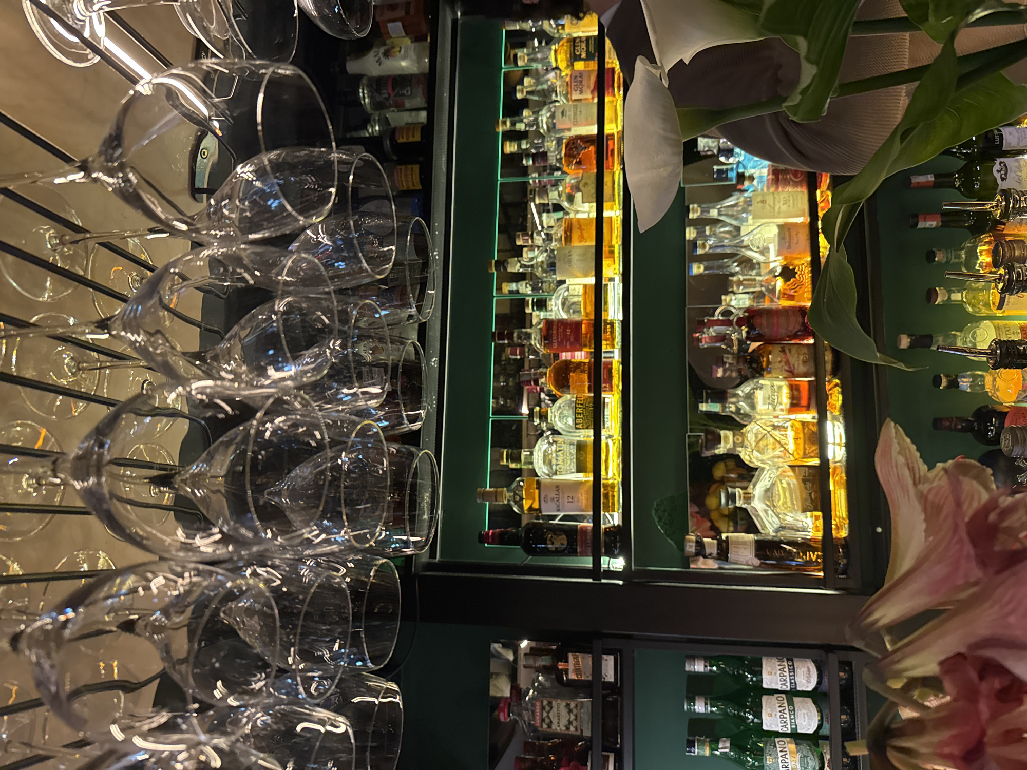 The image, taken in Budva, Montenegro, presents an indoor scene depicting a well-stocked bar or liquor cabinet, likely within a restaurant or lounge. The dominant feature on the left is a rack displaying numerous clean, empty wine glasses of various shapes, hanging upside down, ready for use.
In the center and right, an extensive collection of alcoholic beverages is neatly arranged on dark, possibly green or black, shelves. Many of these bottles are backlit or illuminated from below, casting a warm, golden glow on the spirits. Visible brands include whiskies and scotches such as MACALLAN 12, TOMATIN, ABERFELDY, CLYNELZE, and GLEN MORAY. Other identifiable spirits include LUXARDO AMARETTO and OLIVS.
Further to the right, some wine bottles are visible, a few fitted with pourers. Below these are several green bottles, clearly labeled CARPANO BIANCO and CARPANO CLASSICO, suggesting vermouth or similar aperitifs. Other labels like LUST and Zicape are also present.
Adding a decorative element, parts of a large white flower, resembling a calla lily, and a pink flower, possibly an amaryllis, are visible in the foreground on the right side of the image. The overall lighting suggests an evening or night-time setting, creating an elegant and sophisticated atmosphere for a bar area. No specific activity or event is shown, but the setup implies readiness for service.