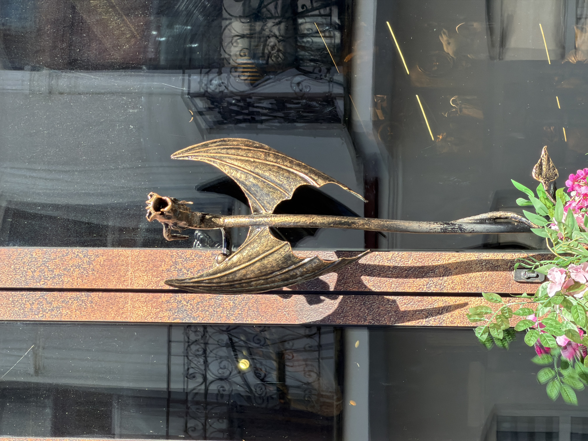 The image shows a bronze dragon door handle on a rustic, possibly rusted, metal door frame. The dragon has its mouth open, and its wings are spread wide. The door handle is positioned vertically, with the dragon's head at the top and its body extending downwards, twisting slightly. Behind the door handle, through a glass window, there are reflections of interior architectural elements, including ornate railings and what appear to be draped curtains. In the foreground, at the bottom of the frame, are artificial pink and white flowers with green leaves. The scene suggests an exterior view, likely a building entrance in Minsk, Belarus, given the prompt's context. The lighting indicates it is daytime, with sunlight casting shadows.