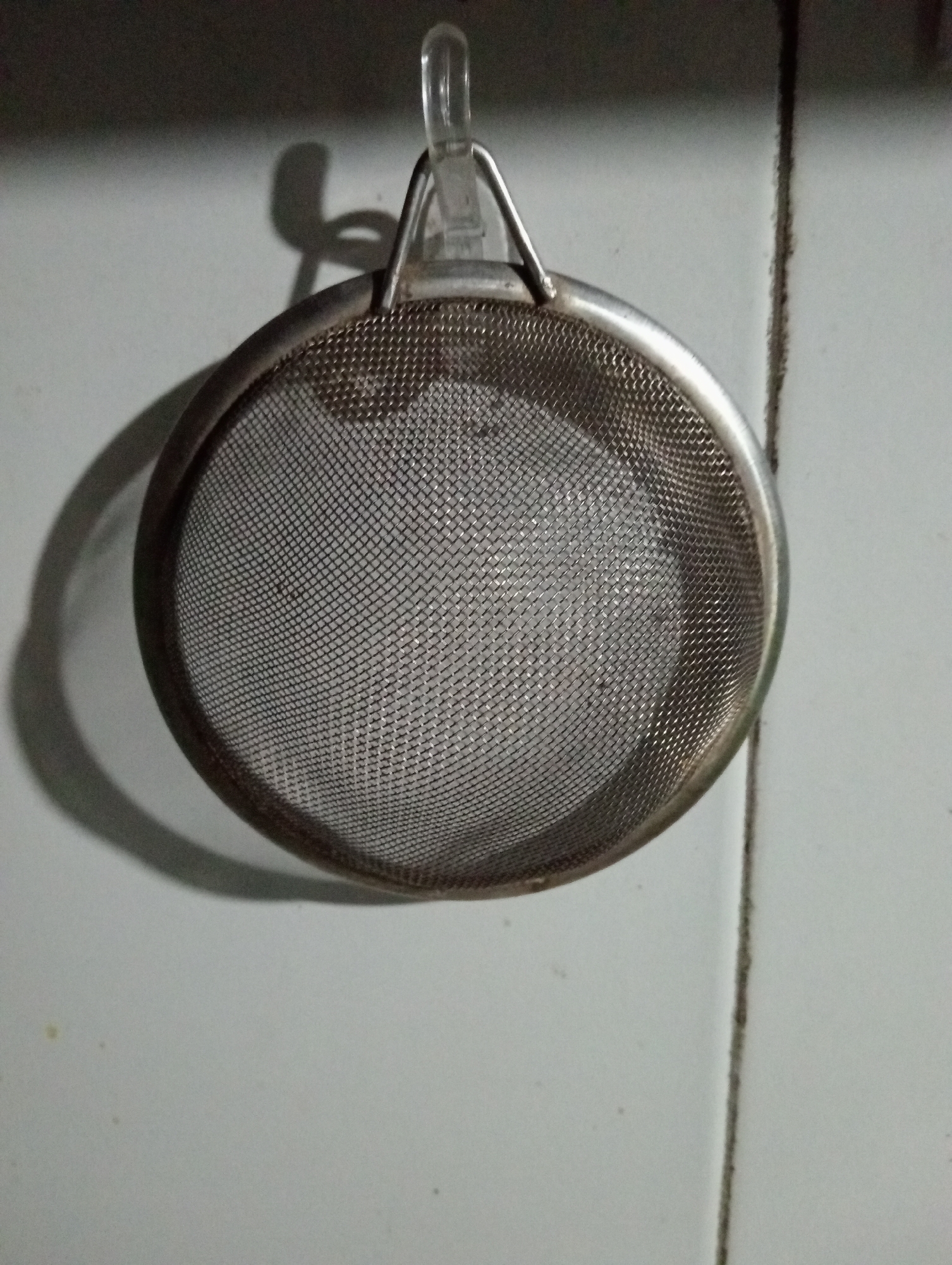 A close-up, eye-level shot shows a metal strainer hanging from a hook on a light-colored tiled wall. The strainer is circular with a fine mesh. A shadow of the strainer is cast on the wall to the left, and a vertical dark line, likely grout between tiles, is visible to the right. The scene is dimly lit, suggesting an indoor setting, possibly a kitchen. The image is devoid of any specific landmarks or contextual clues that would place it within Trenggalek, Indonesia.