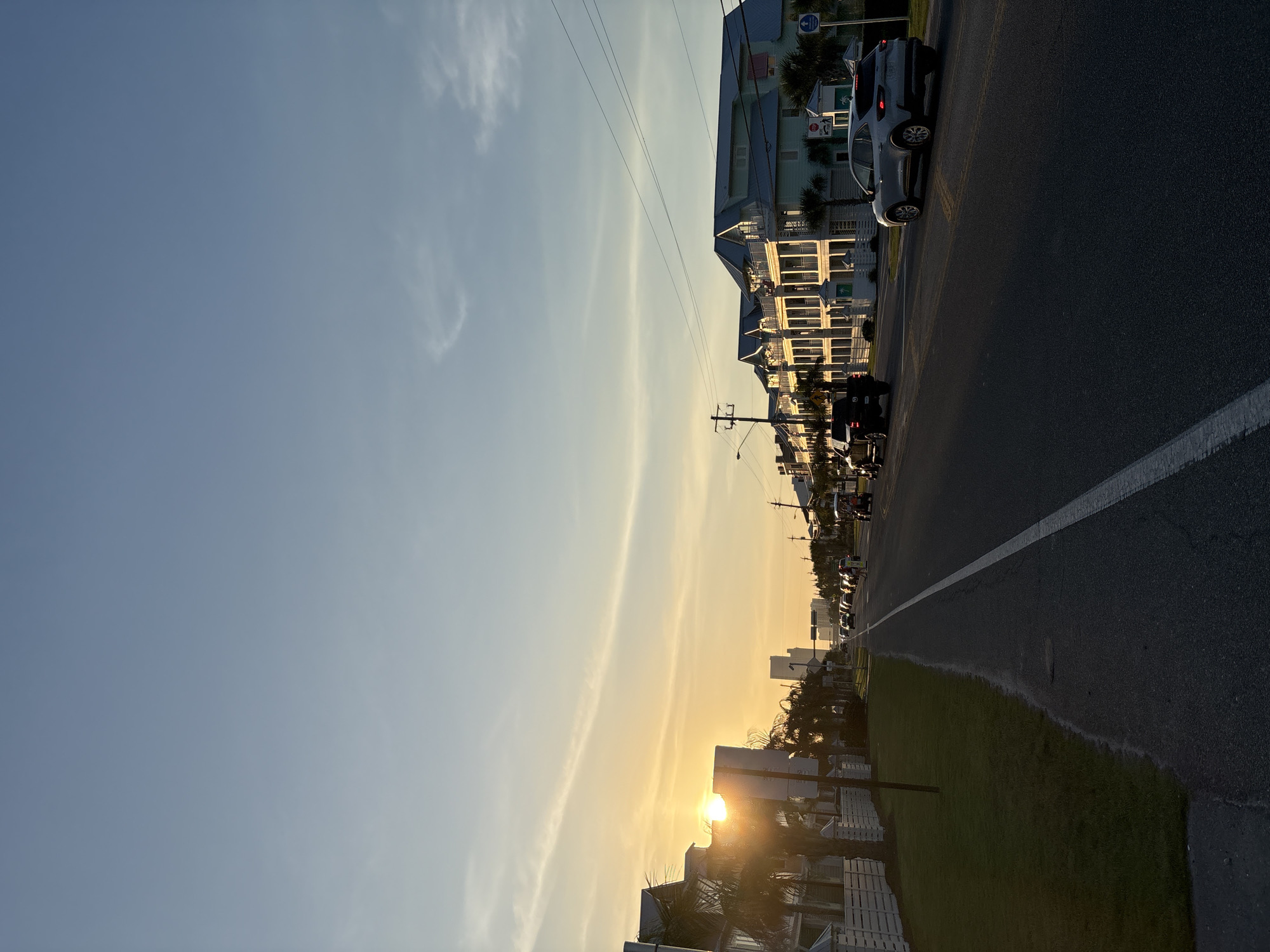 This image captures a street scene in Panama City Beach, United States of America, during the golden hour, likely sunset, given the warm, low-angled light. The sun is a brilliant, bright orb positioned near the horizon on the right side of the frame, casting a golden glow across the landscape and creating long shadows.
The scene features a two-lane asphalt road, visible on the right, with a distinct white solid line marking its edge. A strip of green grass runs parallel to the road, adding a touch of nature to the urban setting. On the left side of the road, a series of multi-story buildings, likely vacation rentals or hotels, line the street, extending into the distance. These structures are predominantly light blue or white, characterized by numerous balconies.
Utility poles with overhead power lines are interspersed along the street. Several vehicles, including a white SUV, are present on the road, either parked or in motion. Palm trees are visible among the buildings and along the roadside, enhancing the coastal resort town feel.
The sky transitions from a soft blue at the top left to a luminous golden and light blue near the setting sun on the right, with a few wispy clouds adding texture. Visible text includes a blue sign on a utility pole on the left displaying "NO PARKING," "NO STOPPING," and a tow-away symbol. Further down, another white sign with a green logo depicting a palm tree is partially visible. The overall impression is one of a serene evening in a bustling coastal destination.