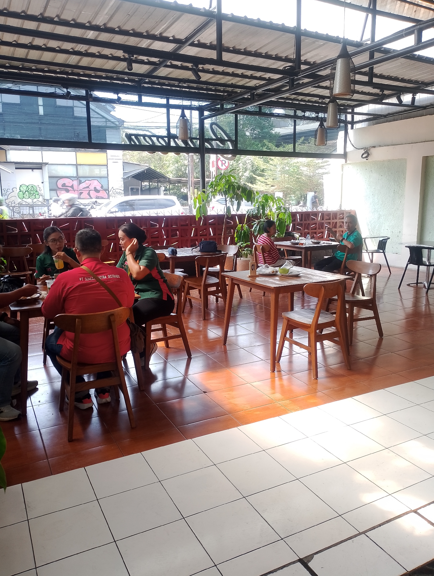The image depicts the interior of a modern cafe or restaurant located in Yogyakarta, Indonesia. The cafe features a high ceiling with exposed metal beams and industrial-style pendant lights. The seating area is furnished with wooden tables and chairs. The floor is a combination of terracotta tiles and white square tiles, with sunlight casting strong shadows from the overhead structure and plants.
In the foreground, four people are seated around a wooden table. One person, wearing a red shirt with "PT SEMESTA INDOSTRA" printed on the back, is facing away from the camera. To their left, two women in green shirts are engaged in conversation, one of whom is drinking from a glass. Another person, partially visible on the far left, is also seated at the table.
In the background, other patrons are seen at different tables. One table has two people, a man in a green shirt and a woman in a striped shirt. Further back, other tables are occupied by individuals or groups, though their details are less clear. Outside the cafe, through large glass windows, trees and buildings are visible, suggesting an urban setting. The overall atmosphere appears relaxed and casual, typical of a daytime gathering at a cafe.