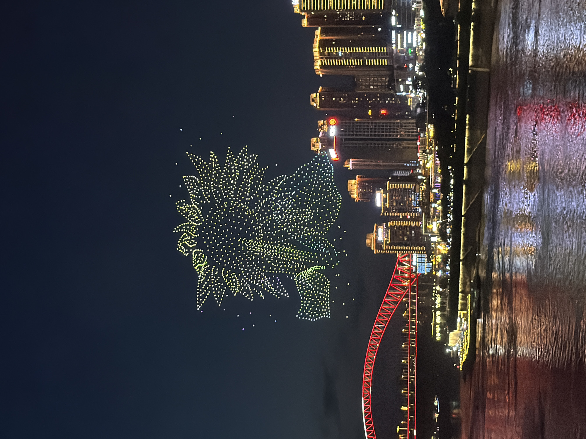 This image captures a nighttime cityscape, likely in Yuzhong District, China, characterized by a drone light show and urban illumination. In the sky, a large, intricate formation of yellow and green lights, resembling a lion's head with a mane, is created by numerous drones. Below this aerial display, a vibrant red bridge arches across the frame, its structure illuminated with crisscrossing lines of light. The bridge appears to span a body of water, where the reflections of the city lights create a shimmering effect on the surface. On the far bank, a row of tall, modern buildings stands lit up, their windows forming grid-like patterns. A large boat, also adorned with yellow lights, is visible on the water near the embankment, further contributing to the illuminated scene. The overall atmosphere is one of festive celebration, with the drone show and city lights creating a visually dynamic spectacle against the dark night sky.
