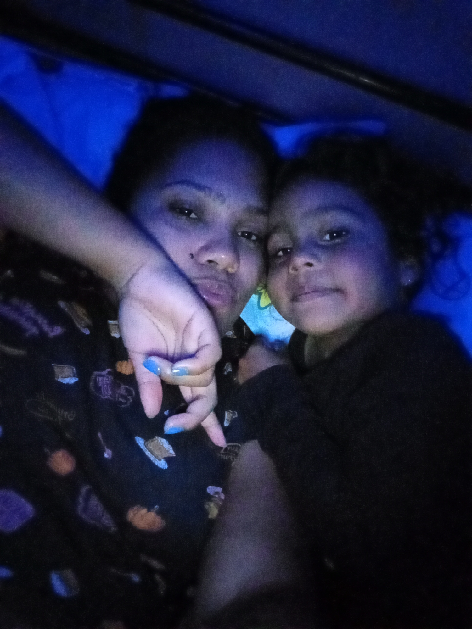 A low-light selfie image depicts two individuals, presumably a woman and a child, under what appears to be blue lighting. The woman has dark hair, fair skin and is wearing a dark patterned shirt with images of cakes and pumpkins on it. Her left hand is in front of her face, displaying light-blue painted nails. The child, with dark curly hair, is positioned close to the woman, with their face partially obscured. The background is dimly lit and includes what appears to be bedding. Based on the prompt, the location is presumed to be Parroquia Ocumare, Venezuela.