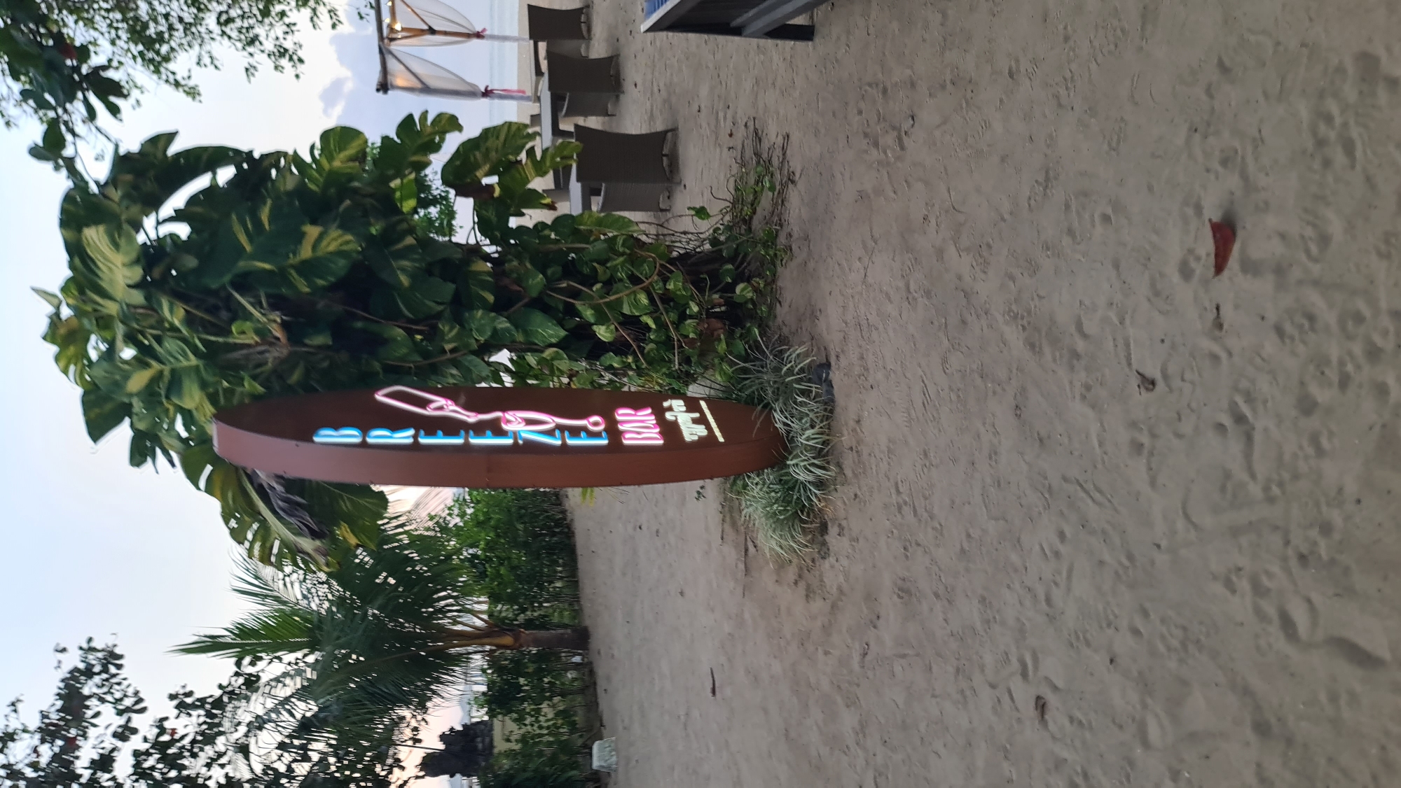 This image depicts an outdoor seating area on a sandy beach, likely located near a bar or restaurant, possibly in Denpasar, Indonesia. A tall, oval-shaped sign with neon lights spelling out "BREEZE BAR" is prominently positioned in the foreground, partially obscured by lush green foliage. The sign is a dark brown color, and the neon letters are illuminated in a vibrant pink and blue hue. The beach is covered in light brown sand, showing footprints. In the background, several tables and chairs are set up under a canopy structure, suggesting an area for patrons to relax and enjoy drinks or meals. The ocean is visible in the far background, with gentle waves lapping at the shore under a hazy sky. Tropical trees and palm trees surround the area, creating a serene and relaxing atmosphere.