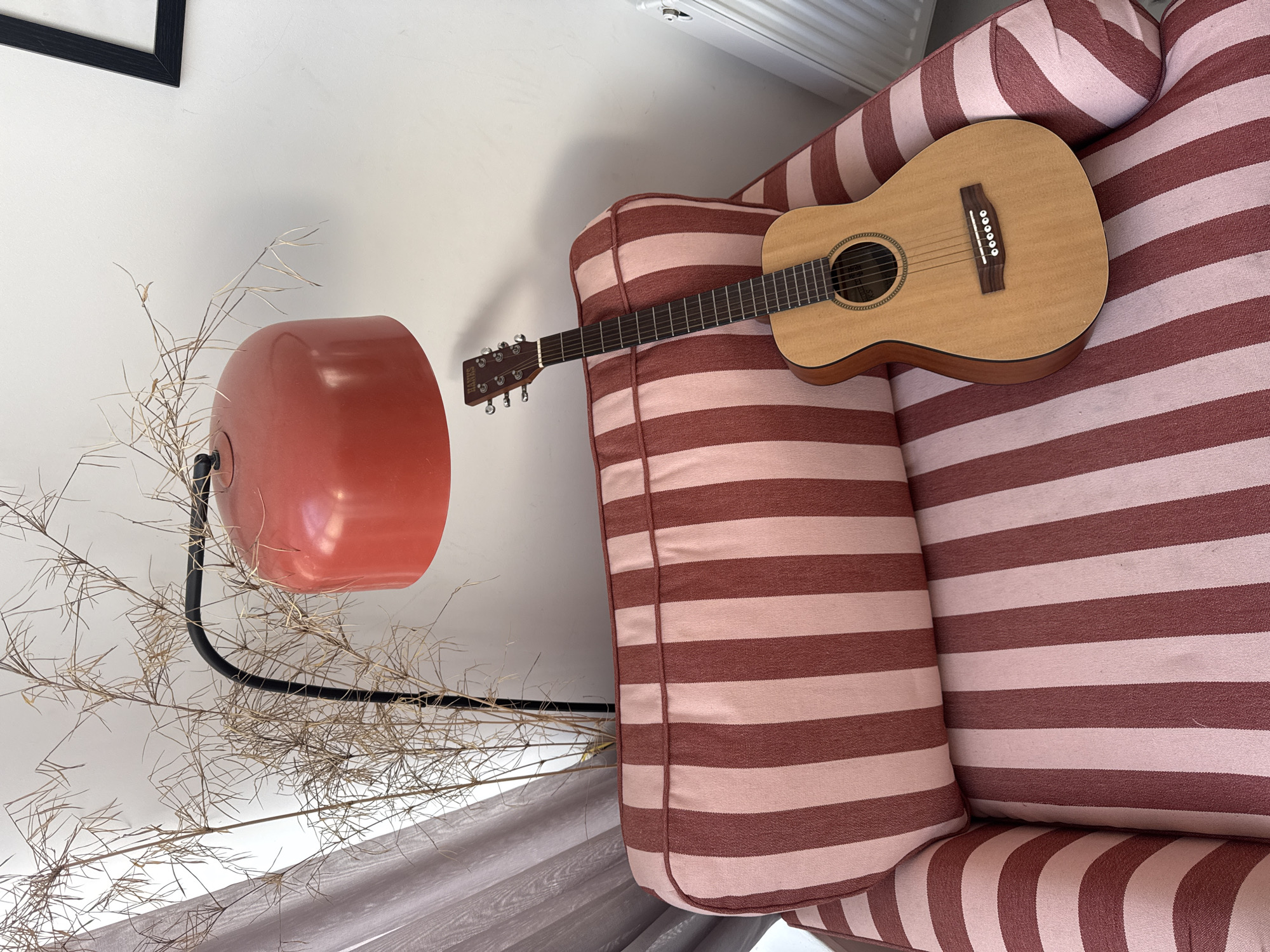 A medium, overhead shot shows a light-brown HAYS guitar lying diagonally on a pink and red striped couch, positioned next to a red lamp with a black stand, around which are arranged tall, dry plants. A partial black frame is visible in the upper left. The scene is indoors, with the floor partially visible at the bottom. The location is not identifiable as being in London, United Kingdom.