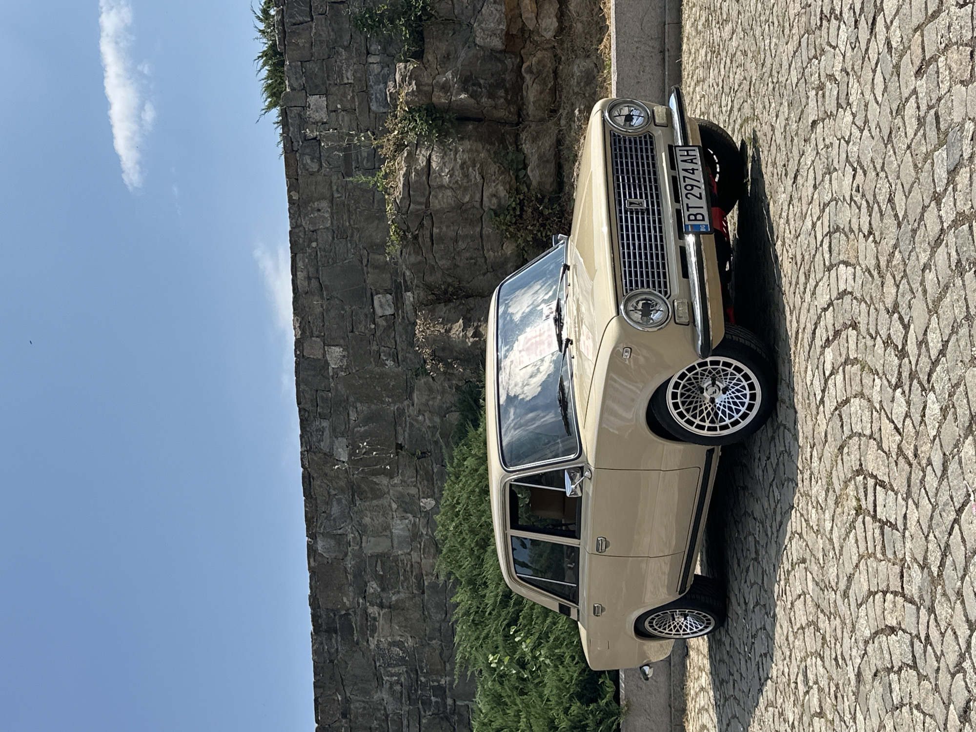 A vintage, beige sedan, identified by its Bulgarian license plate "BT 2974 AH", is parked on a cobblestone surface in front of a rough-hewn stone wall. The car, a Lada 2106, has chrome accents on its grille, headlights, door handles, and mirrors. Its interior appears to be tan. Lush green bushes are visible behind the car, partially obscuring the lower portion of the stone wall. Above the wall, a clear blue sky with scattered white clouds dominates the upper part of the image. The scene is bathed in sunlight, casting shadows from the car onto the cobblestones. This setting is likely the Tsarevets fortress in Veliko Tarnovo, Bulgaria, a popular historical site known for its ancient stone architecture and cobblestone pathways. There are no people or animals present in the frame.