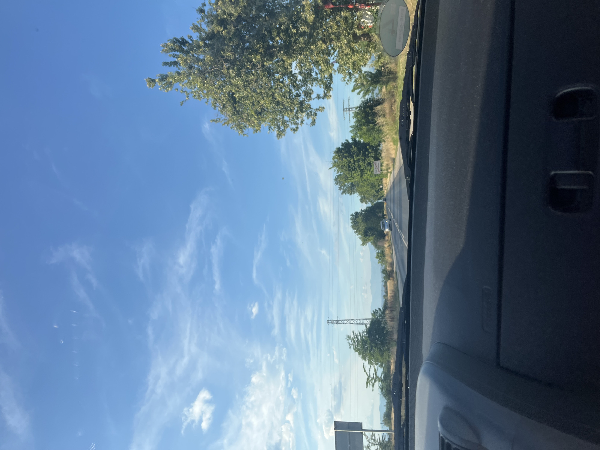 The image is taken from the interior of a vehicle looking forward towards a two-lane road. The sky is bright blue with scattered white clouds. Trees line both sides of the road, with a large tree dominating the right side of the frame. In the distance, power lines and pylons are visible. The road appears to be in a rural area, with fields and hills in the background. The dashboard of the vehicle is visible in the foreground. The image appears to have been taken in or around Graf Ignatievo, Bulgaria, given the presence of a round sticker with Cyrillic writing on the windshield, typical of vehicles in Bulgaria.
