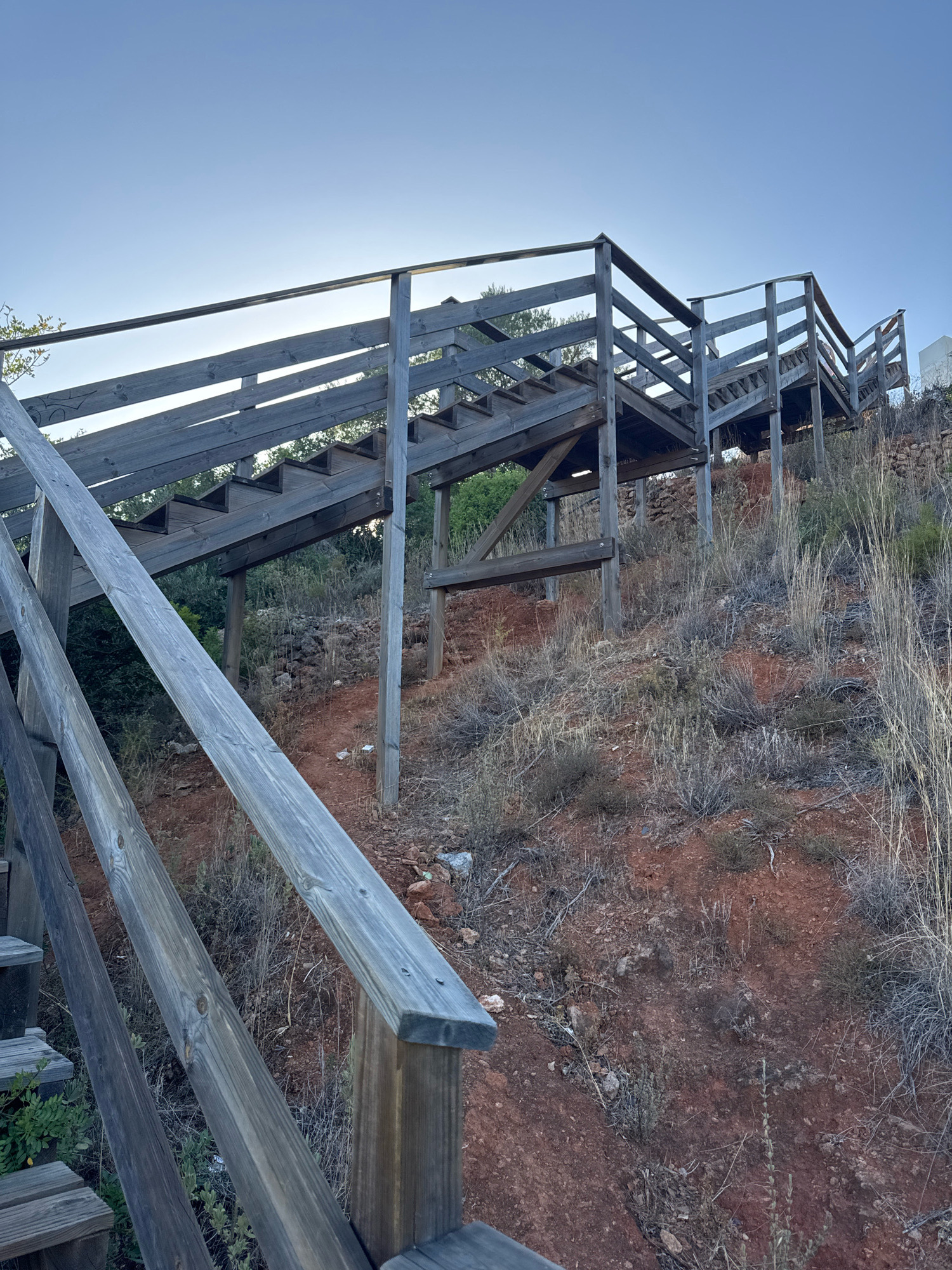 An outdoor wooden staircase ascends a dry, reddish-brown hillside covered in sparse, dry vegetation and rocks. The staircase is constructed from weathered gray wood, featuring railings and multiple flights of steps. The sky above is a clear, pale blue. The scene appears to be located in a natural or semi-natural environment on the outskirts of Loulé, Portugal, possibly a hiking trail or access to a viewpoint. There are no people visible in the image.